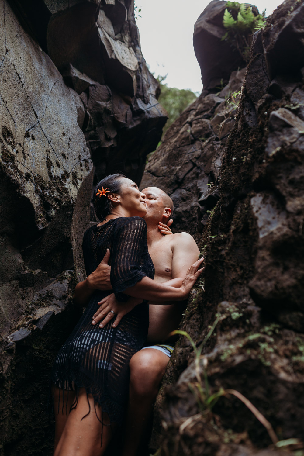 Couple embraces as he kisses her cheek leaned up against rocks during their Kauai engagement photoshoot by Liz Koston.