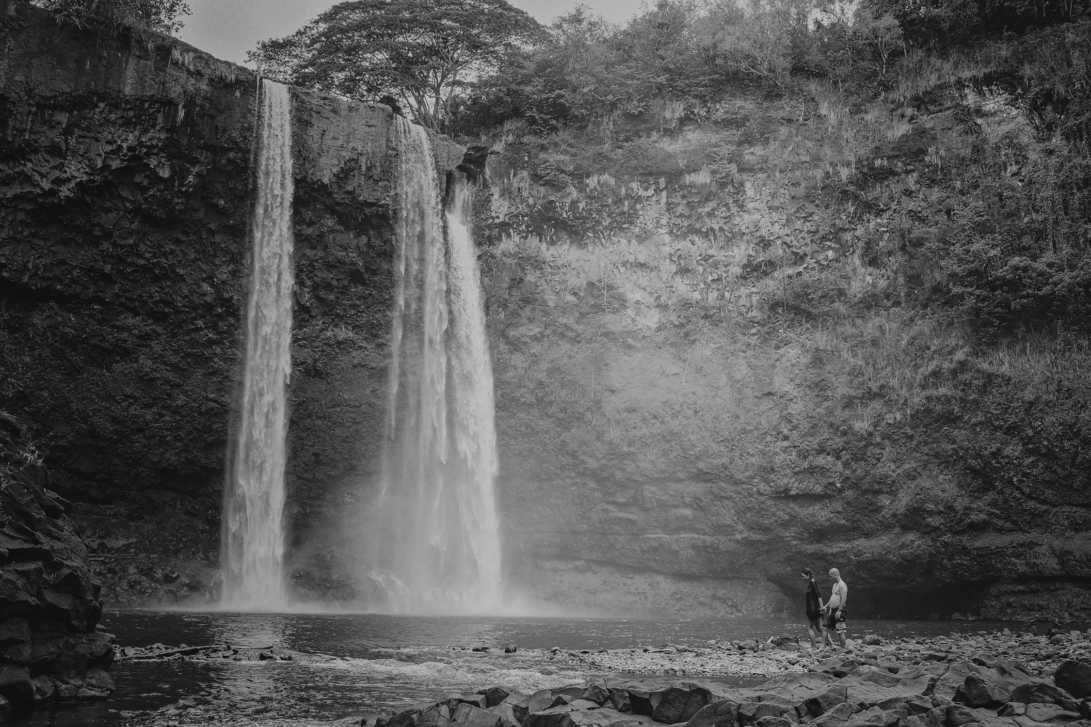 Couple stands at the base of a large waterfall in Hawaii during their Hawaii engagement photoshoot by Liz Koston.