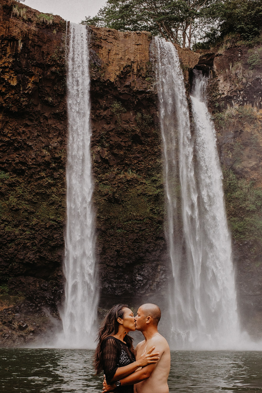Couple kisses at the base of a large water during their couples engagement photoshoot in Hawaii by Liz Koston.