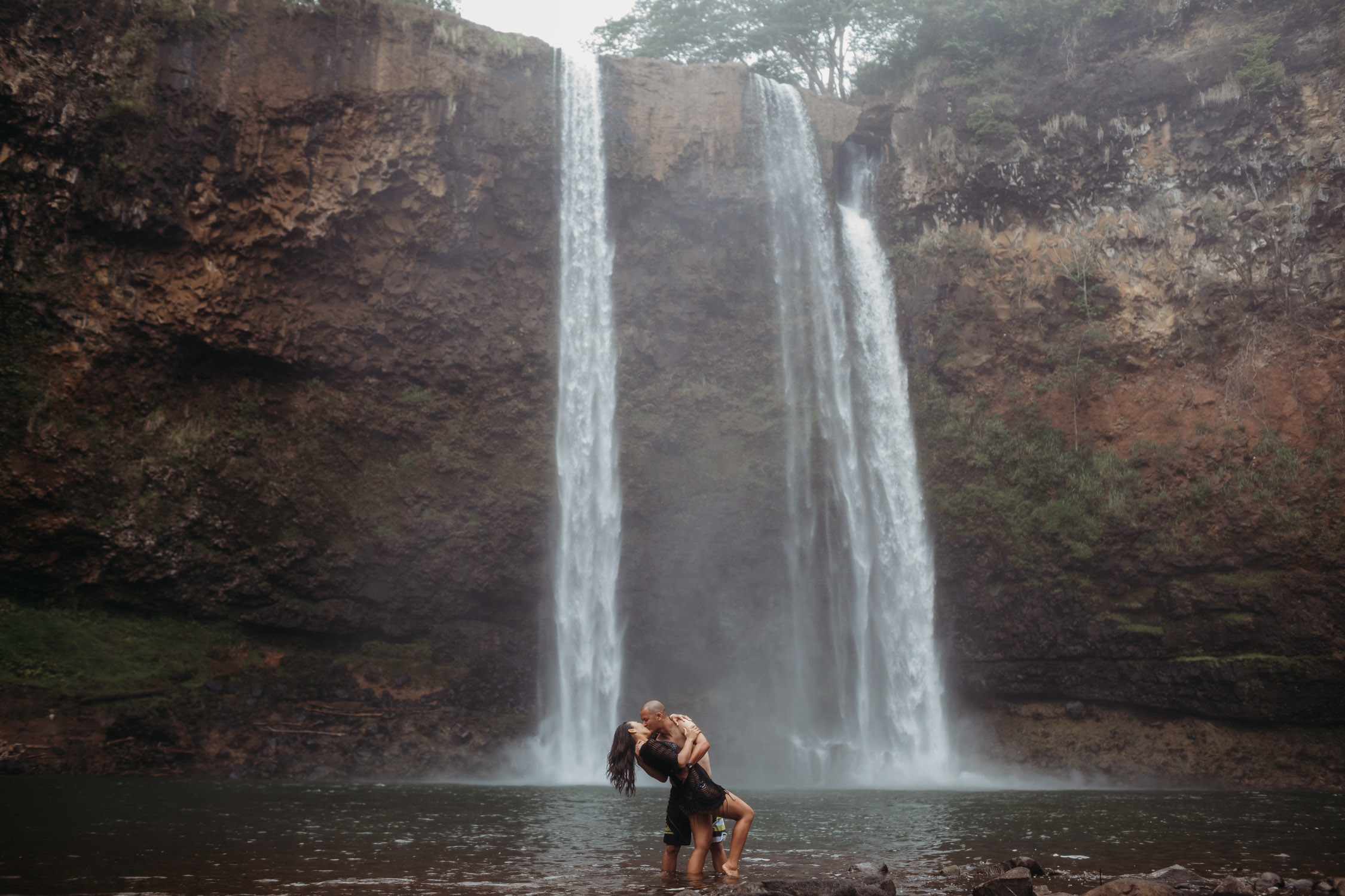 Couple kisses in front of a large waterfall during their Hawaii engagement photoshoot by Liz Koston.