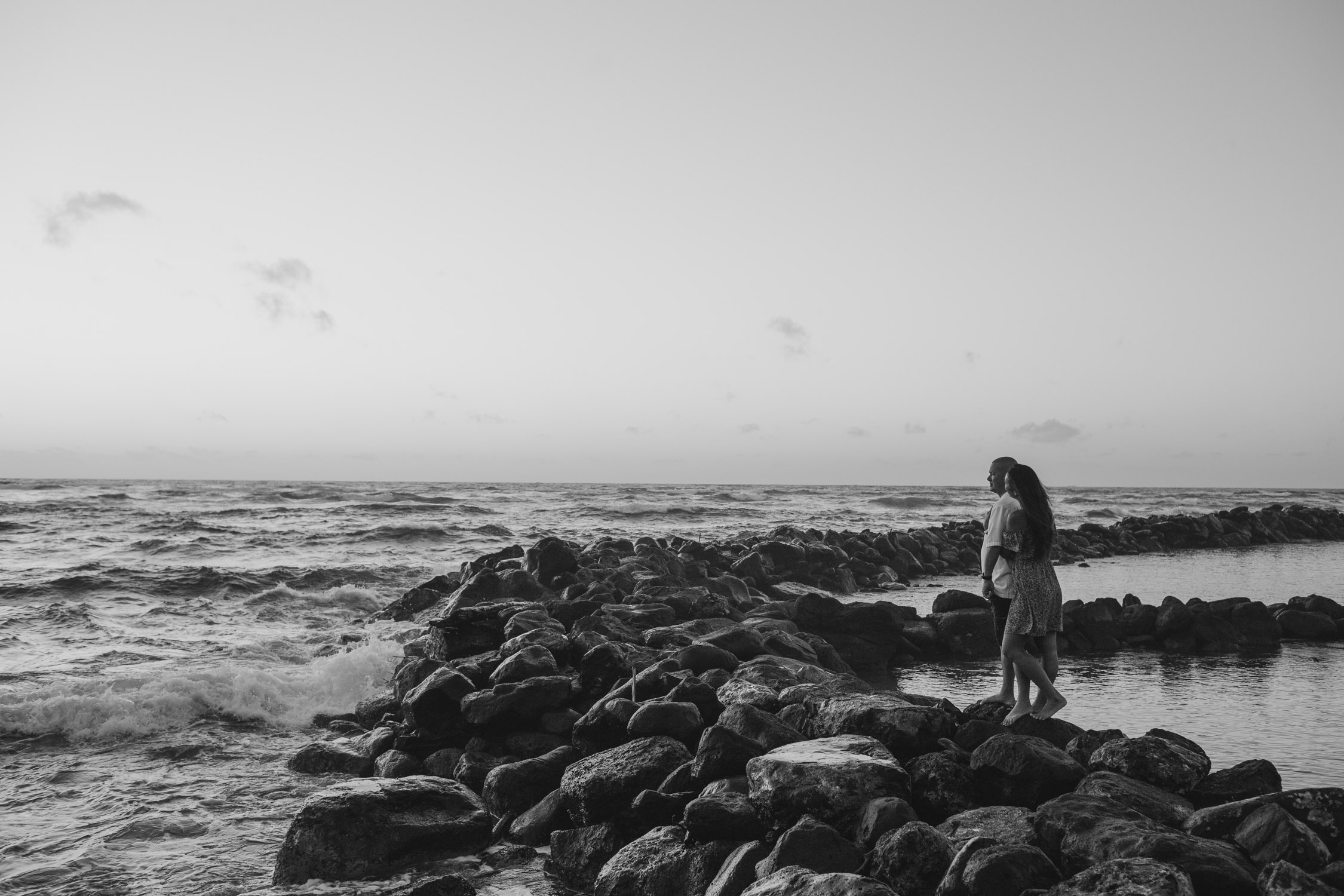 Couple stands on rocks with waves crashing around them during their Hawaii engagement photoshoot by Liz Koston. 