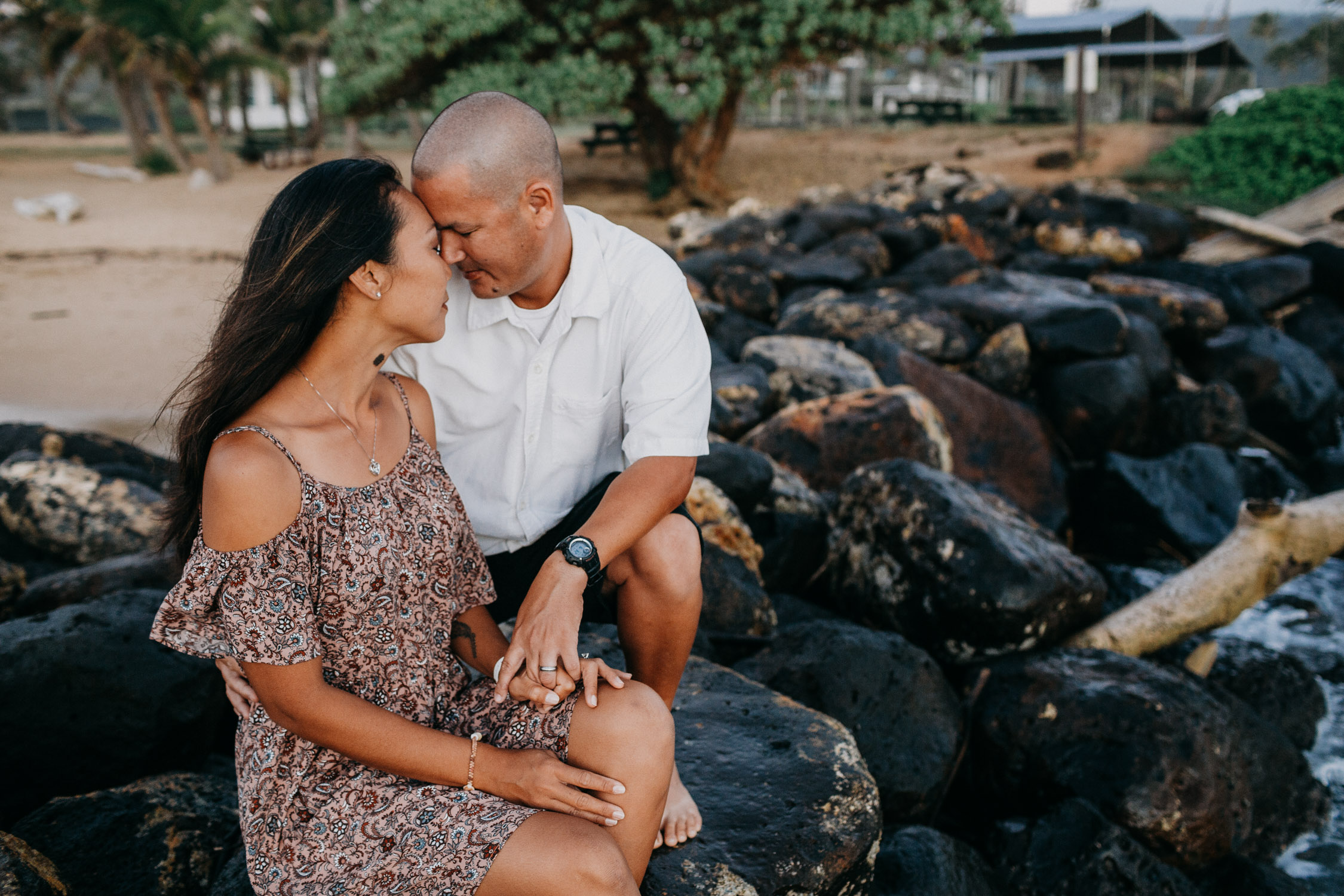 Couple touches foreheads while sitting on rocks overlooking the beach in Kauai, Hawaii.