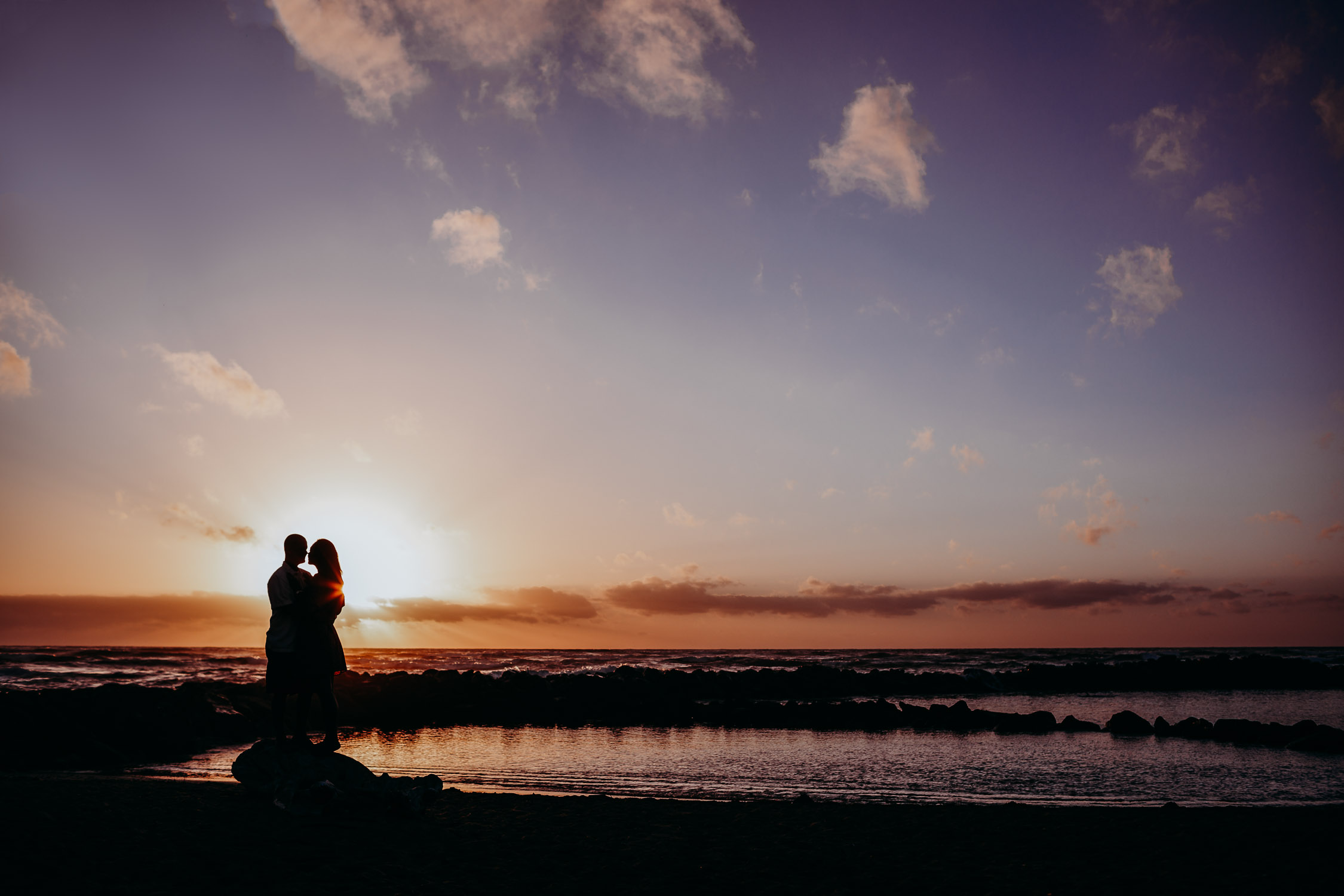 Couple stands in an embrace as they watch the sunset during their Kauai engagement photoshoot by Liz Koston.