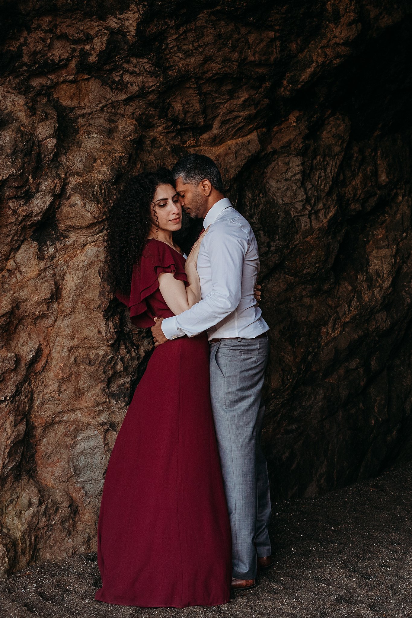 Couple stands against a rocky beach wall in a loving embrace during their Sutro Baths engagement photoshoot