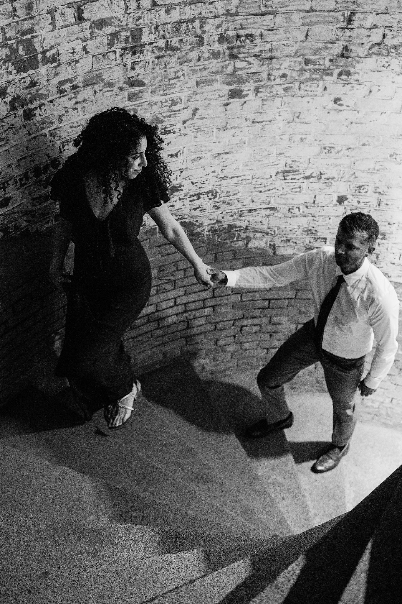 Black and white image of woman leading her fiance up a spiral staircase during their Fort Point engagement photoshoot.