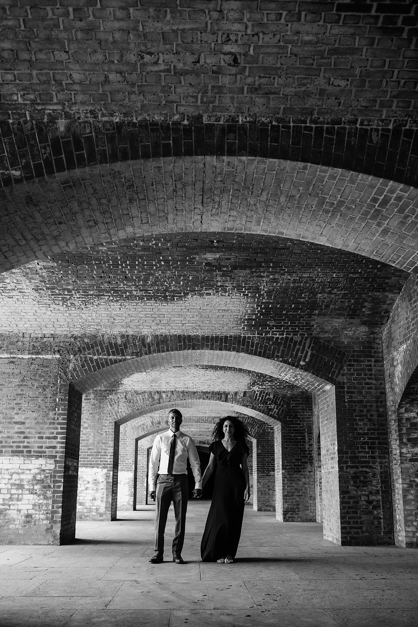 Black and white image of couple facing the camera holding hands in a brick tunnel at Fort Point in San Francisco, CA