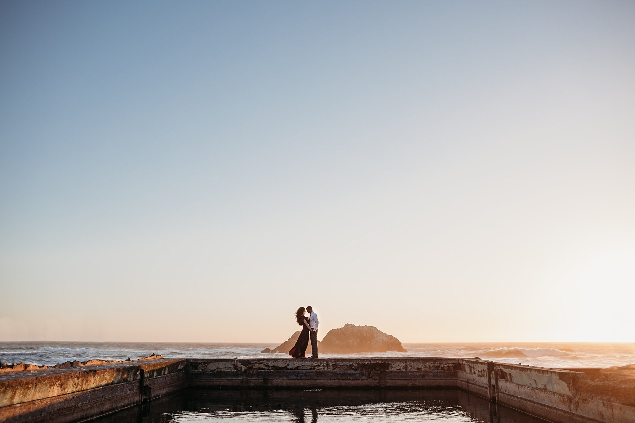 Couple stands on the ledge at Sutro Baths pool at sunset during their San Francisco engagement photoshoot.