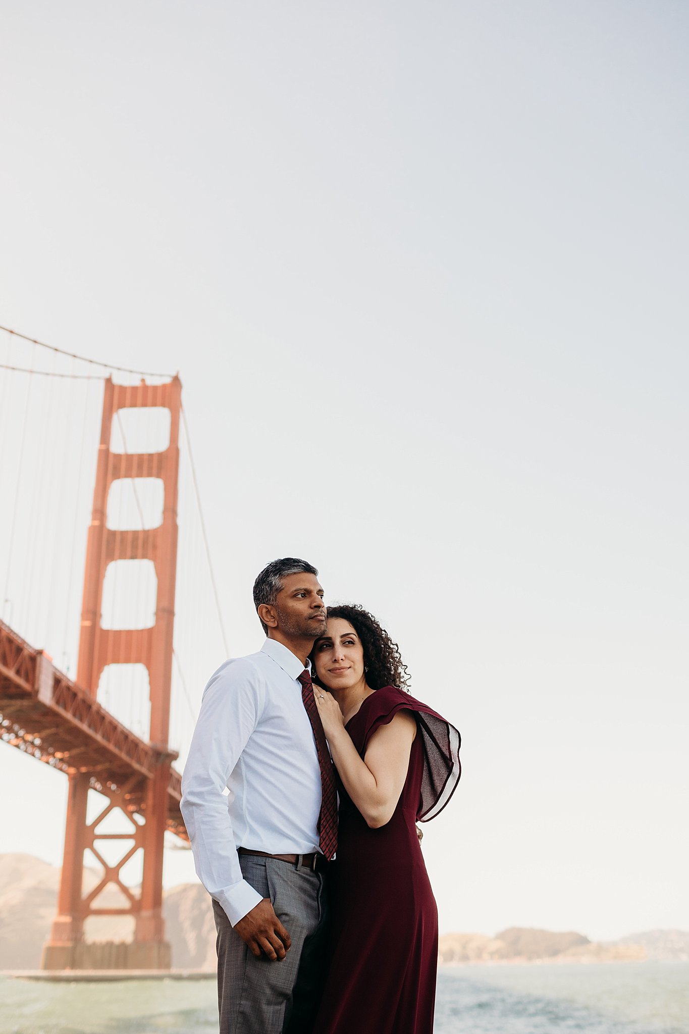 Woman in flowy red dress rests her head on her fiance's shoulder underneath the Golden Gate Bridge at Fort Point, San Francisco