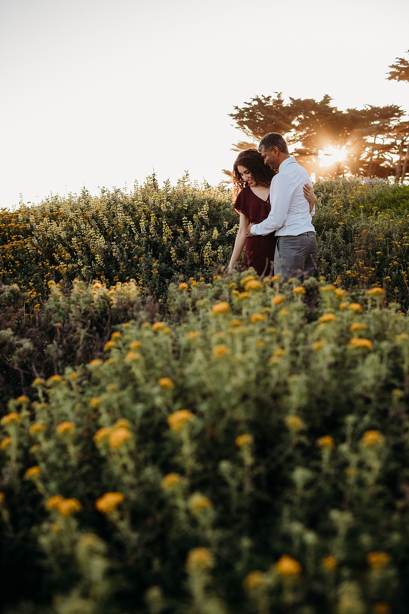 Couple stands in a field of yellow flowers at sunset during their San Francisco engagement photoshoot.