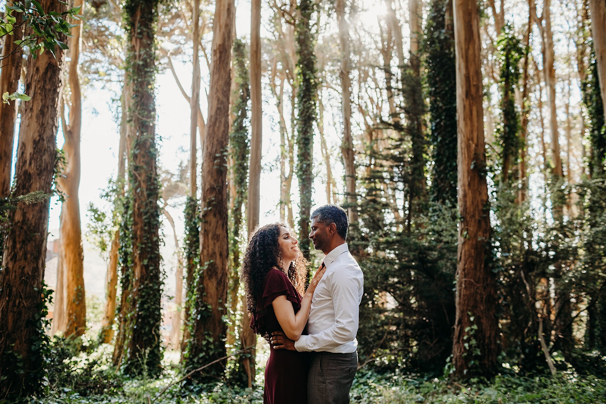 Smiling couple stands in an embrace amongst tall trees during their engagement photoshoot at Lover's Lane in San Francisco. 