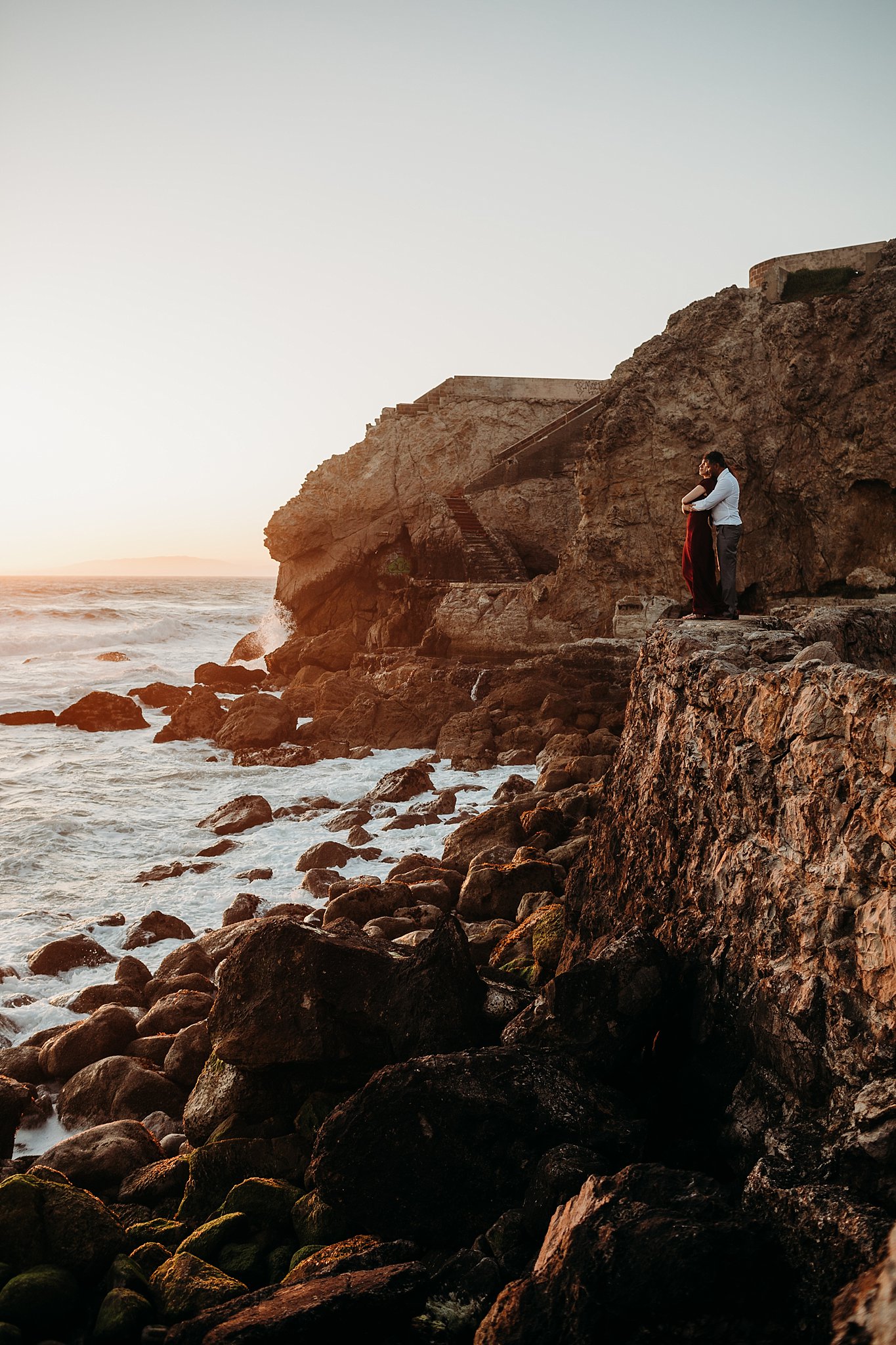 Couple stands on a rocky ledge overlooking the ocean at Sutro Baths during their sunset engagement session in San Francisco, CA