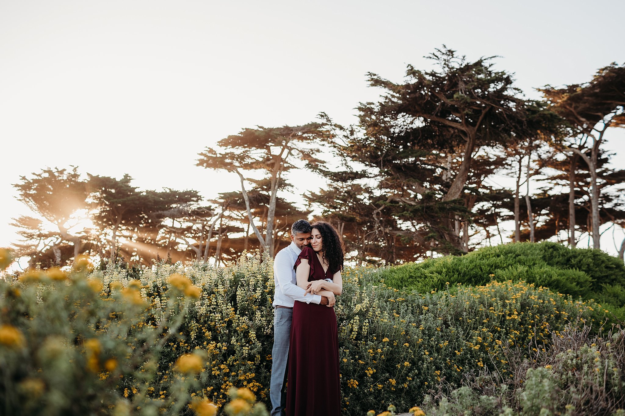 Man hugs woman from behind while standing in a field of yellow flowers at Sutro Baths during their engagement photoshoot. 