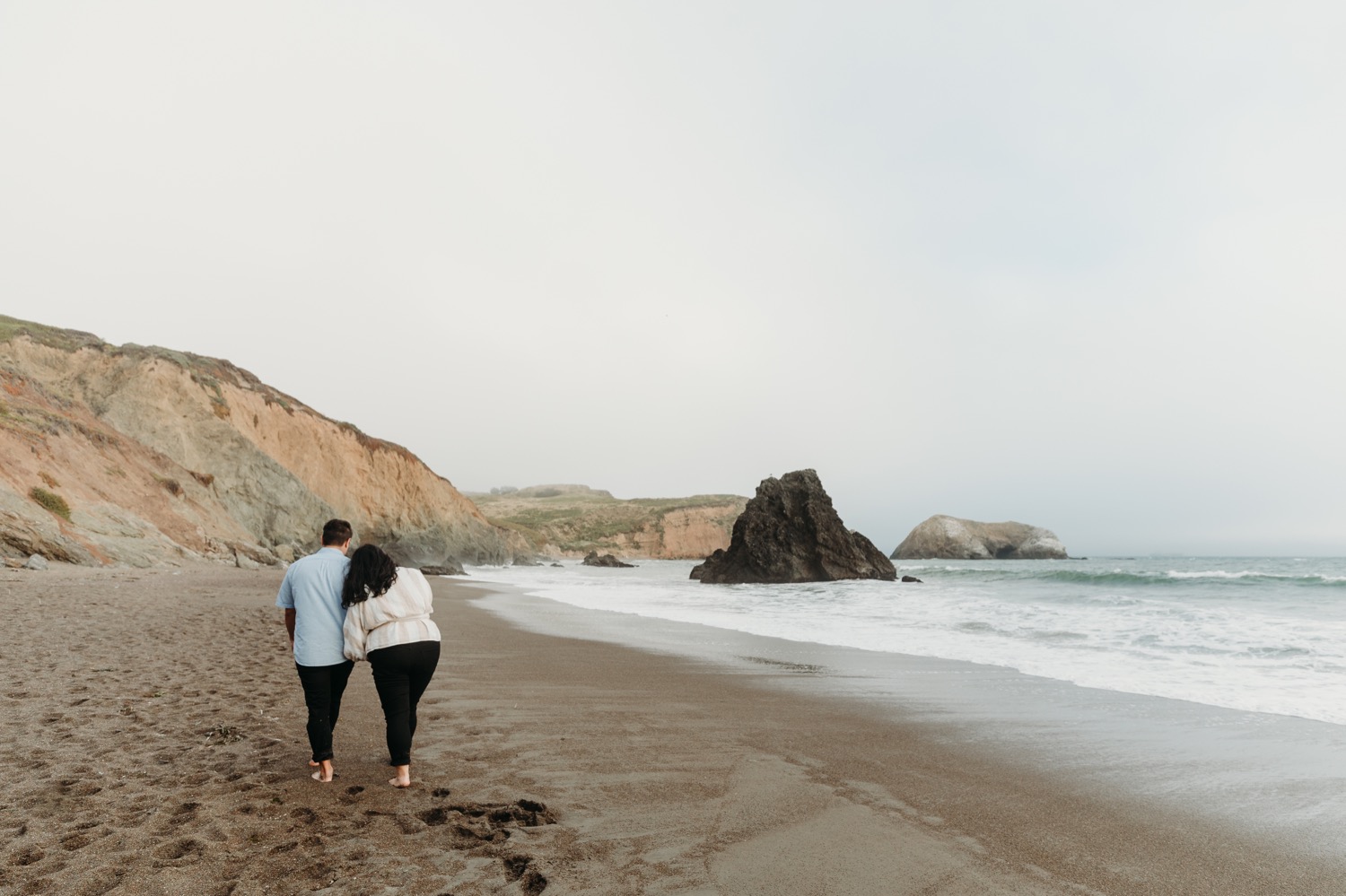 Couple walks away from the camera on the sandy beach at Rodeo Beach, CA during their engagement photoshoot.