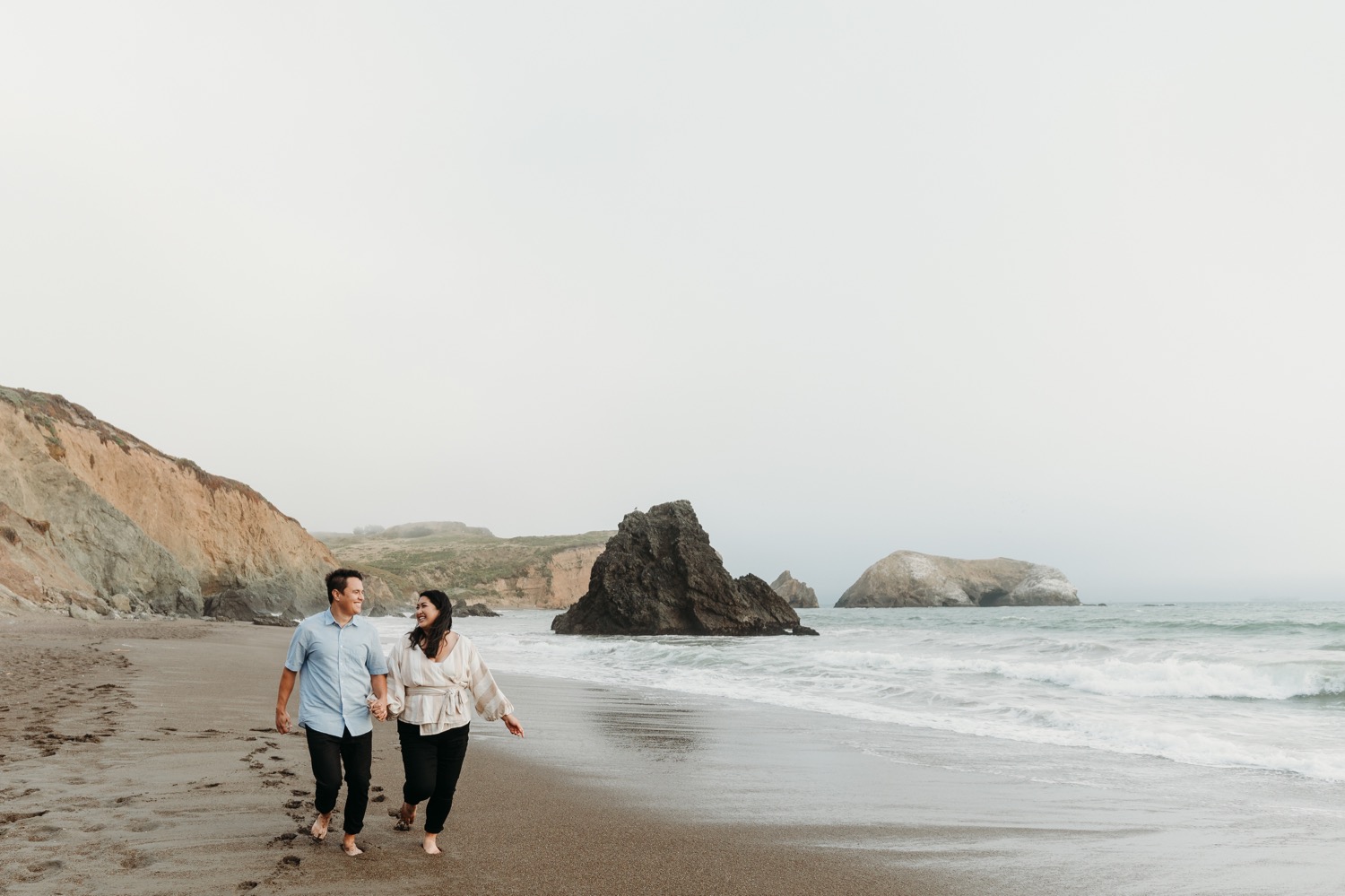 Smiling couple walks hand in hand on the sand during their Rodeo Beach engagement photoshoot.