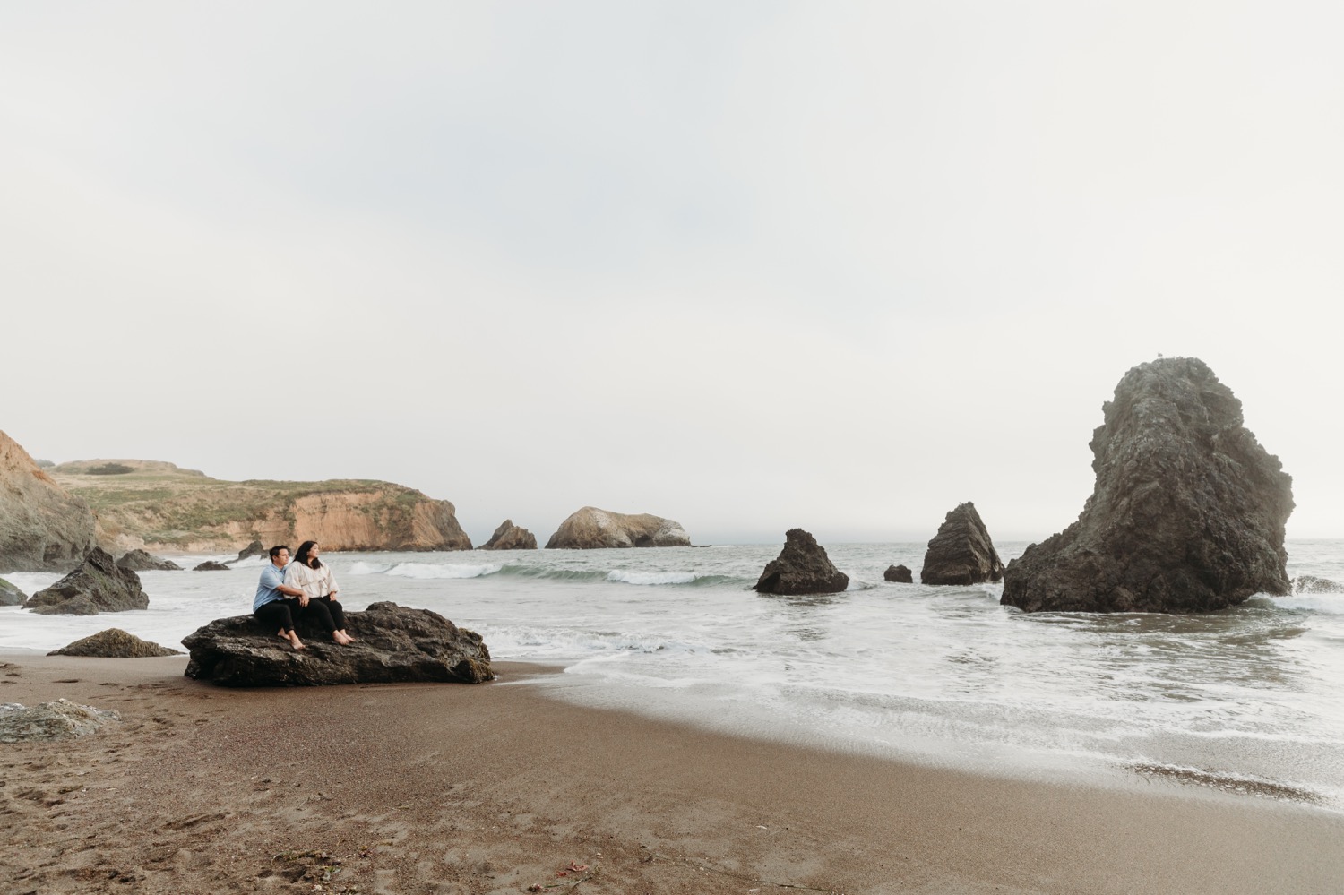 Couple sits on rock on the gazing at the Rodeo Beach rock stack during their engagement photoshoot.