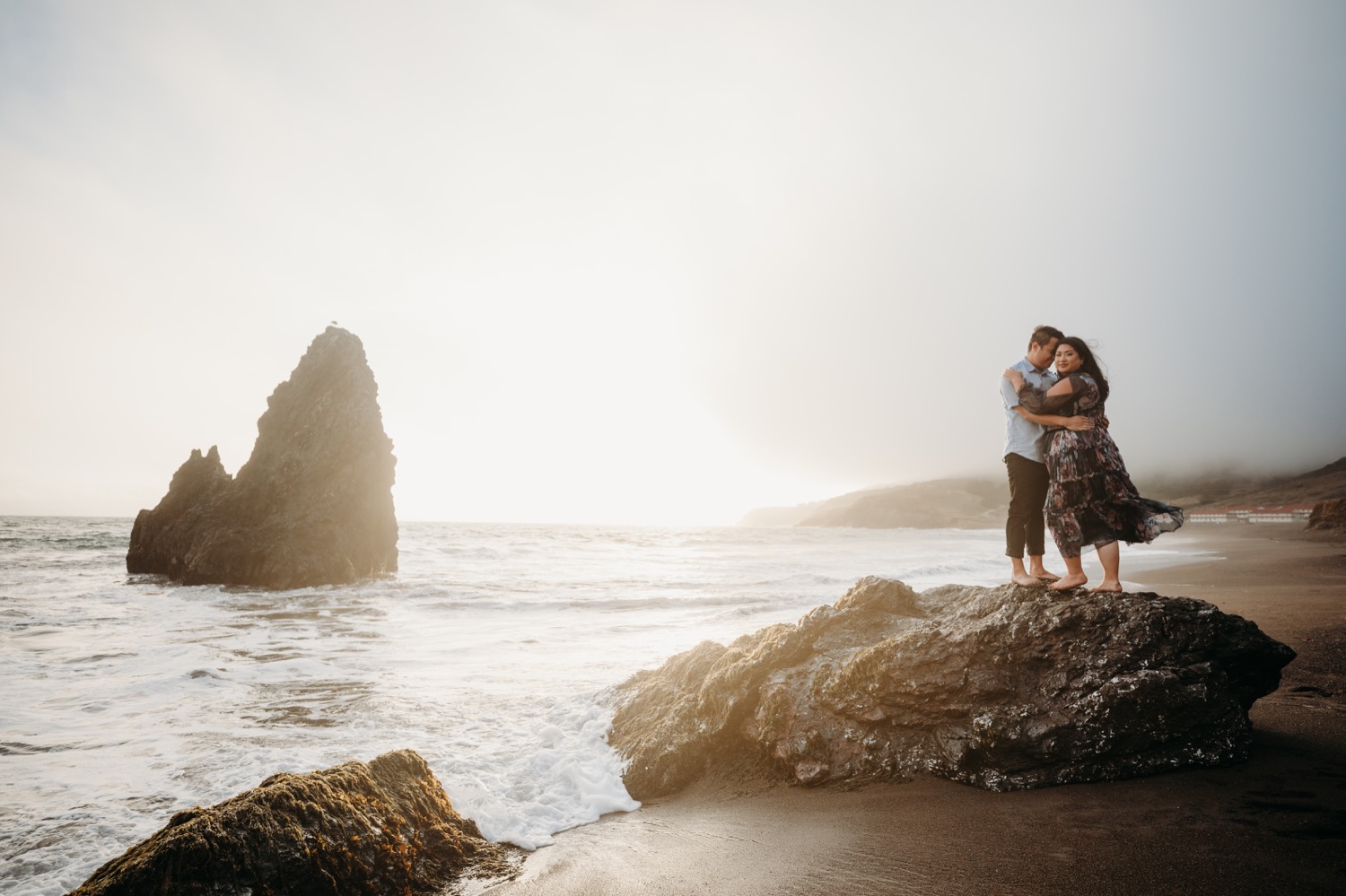 Couple stands on rock at Rodeo Beach at sunset as the wind blows the womans flowing dress and the man nuzzles her during their Rodeo Beach engagement photoshoot.