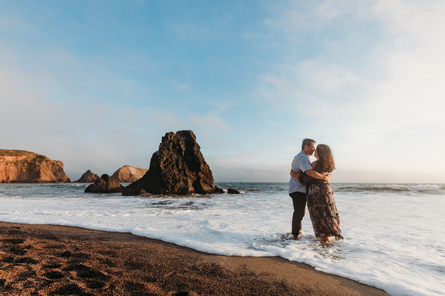 Couple kisses while standing ankle deep in the waves during their Rodeo Beach sunset engagement photoshoot.