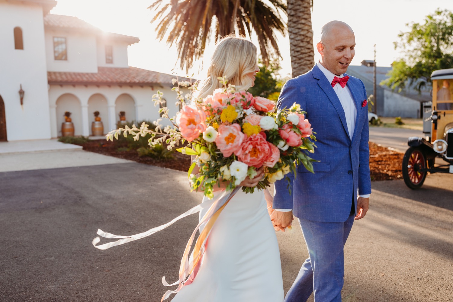 Bride holding her spring colored bouquet walking with her groom during sunset. Spanish style building in the back.