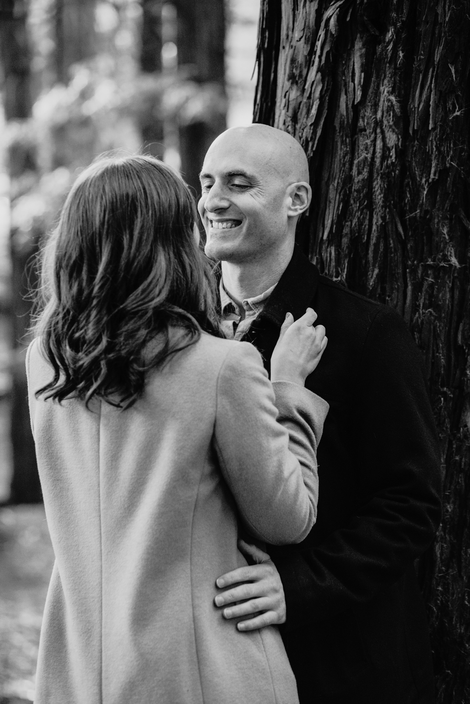 Man leans against a redwood tree as woman faces him and they hold each other during their Golden Gate Park engagement photoshoot.