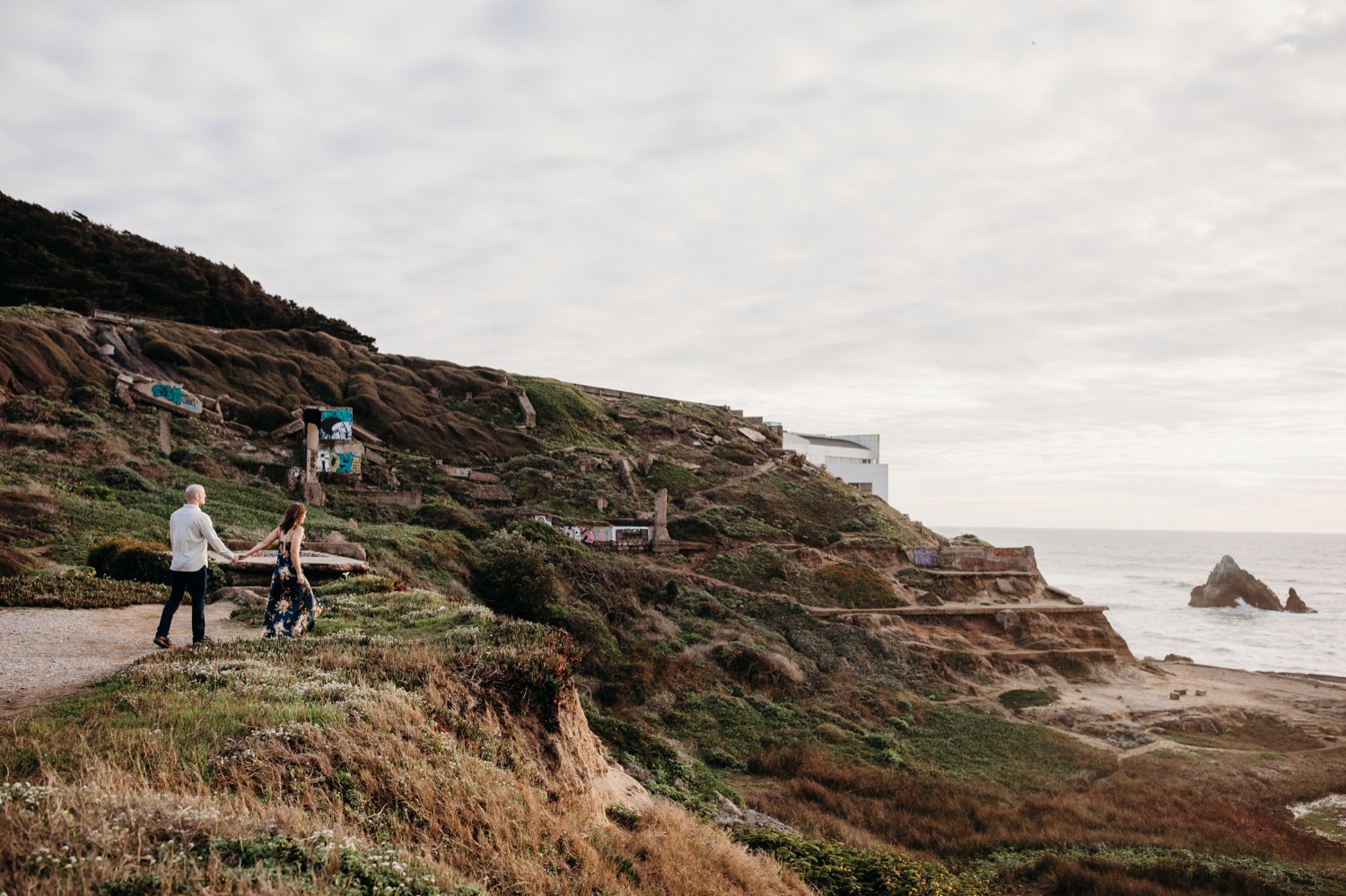 Woman leads her fiance down a path on their Sutro Baths engagement photoshoot.