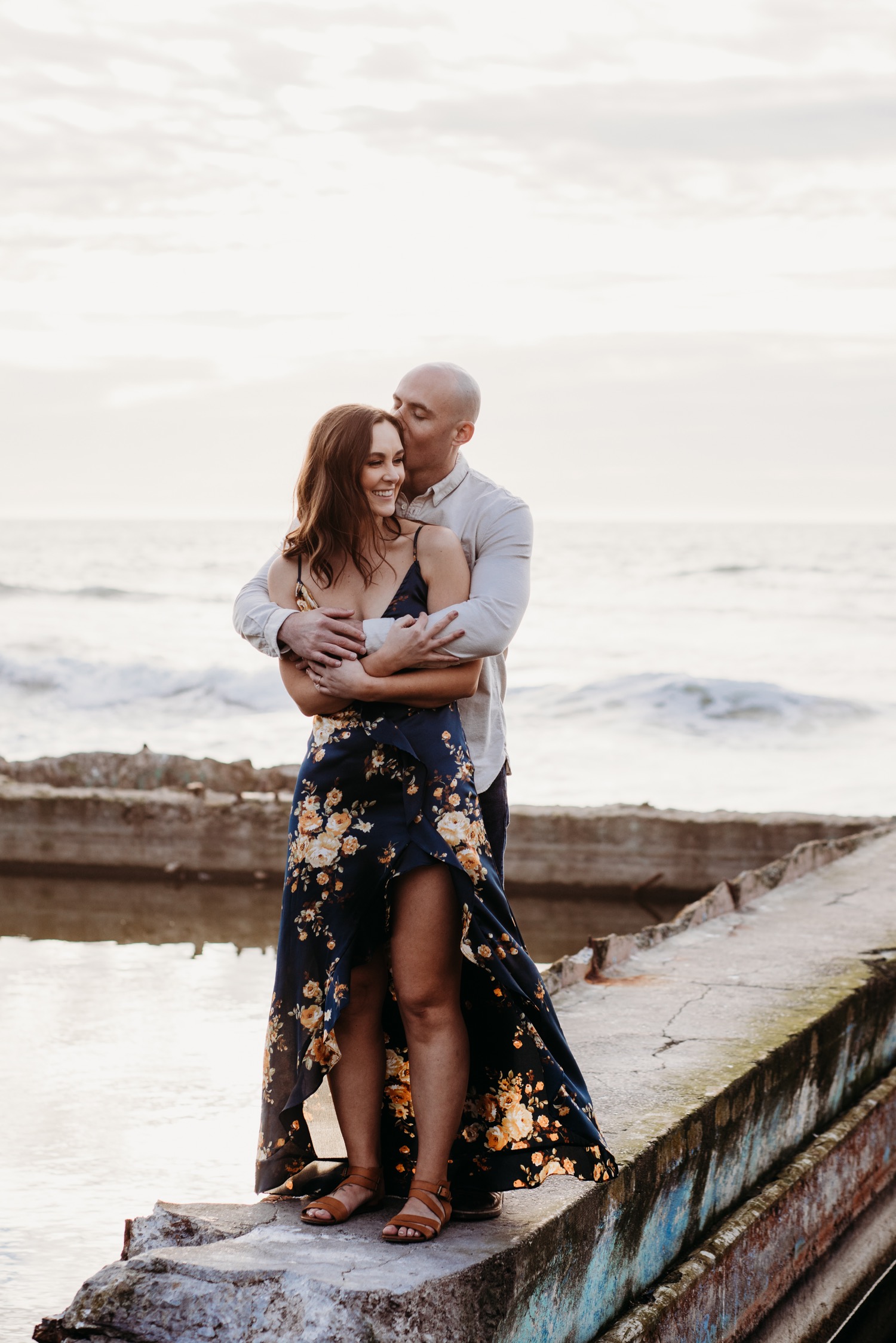 Man hugs his fiance from behind while standing on a Sutro Bath ruin and the Pacific Ocean in the background during their Sutro Baths engagement photoshoot.