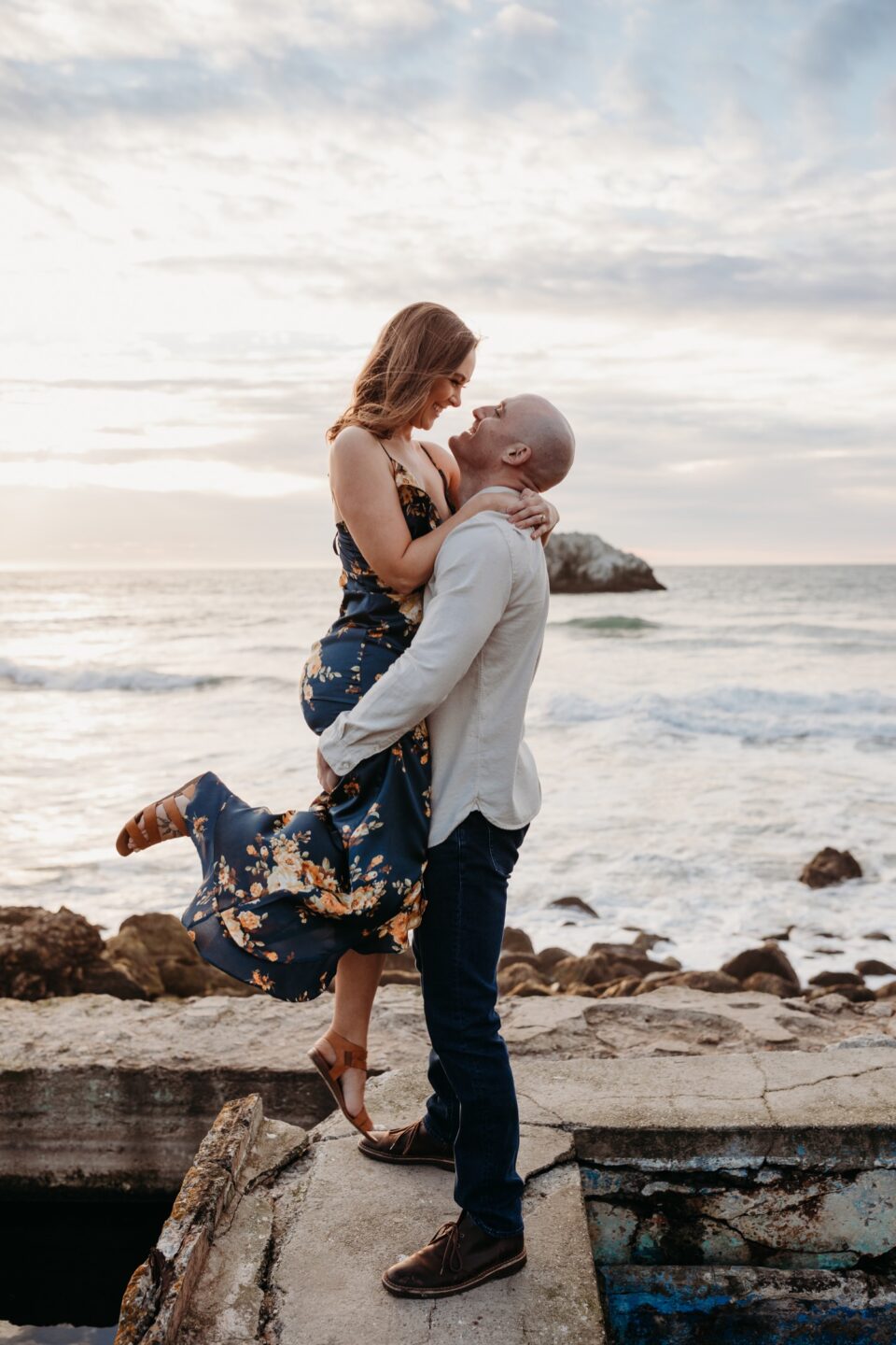 Man picks fiance up during their sunset engagement photoshoot at Sutro Baths in San Francisco.