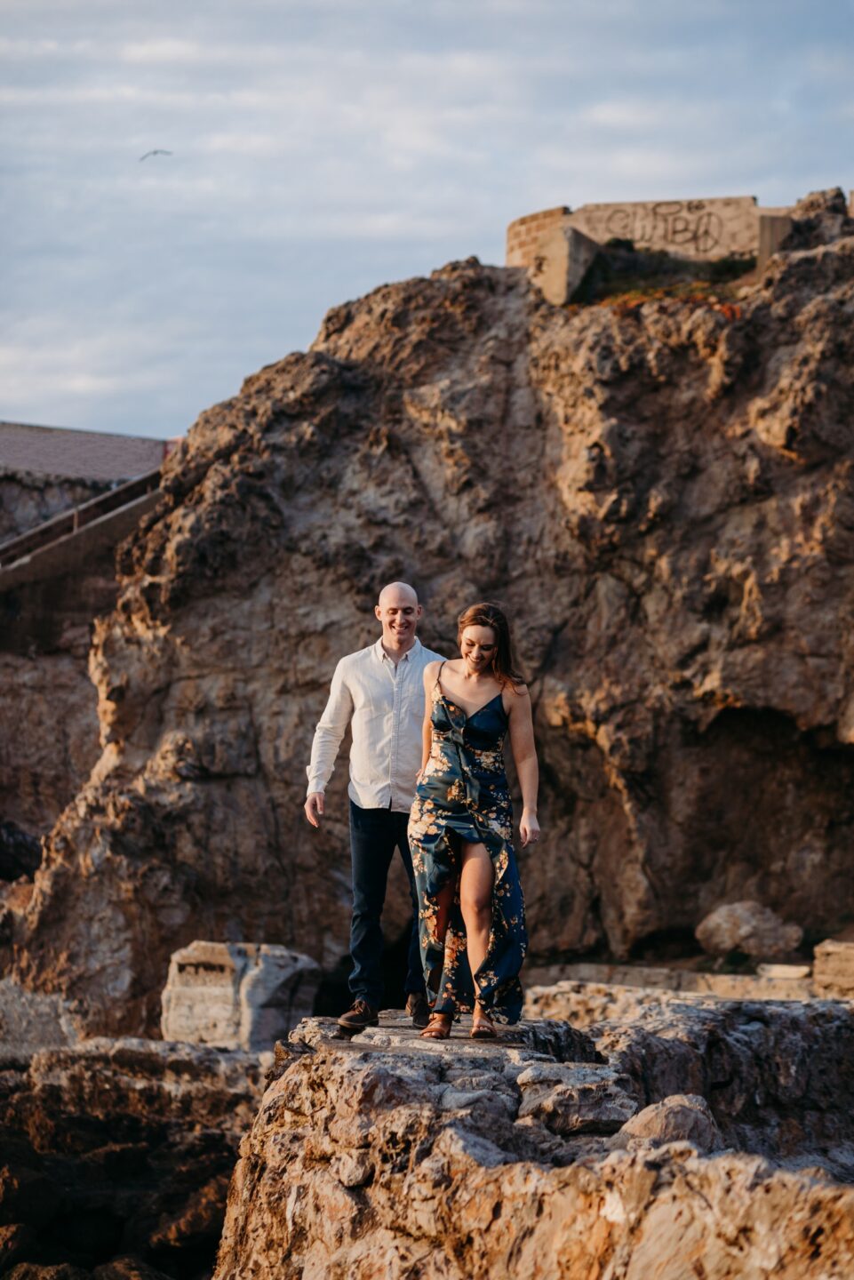 Woman leads her fiance on a rocky path during their Sutro Baths engagement photoshoot.