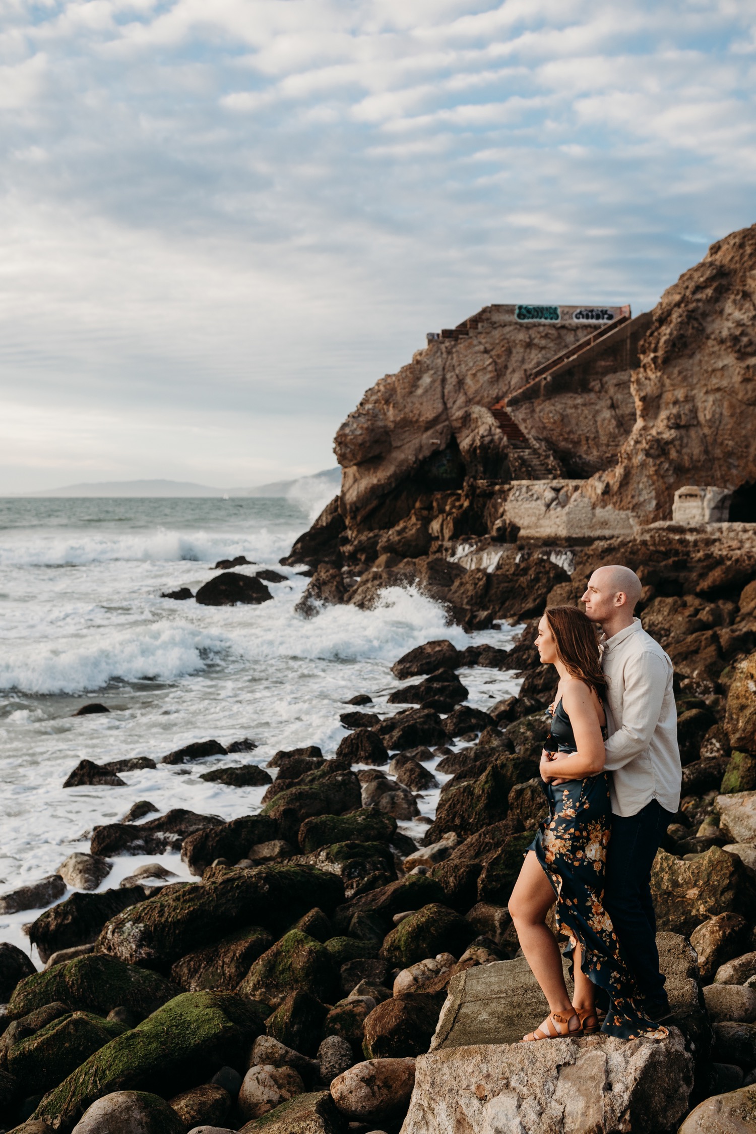 Couple stands on rocky beach while gazing at the Pacific Ocean during their sunset engagement photoshoot in San Francisco.