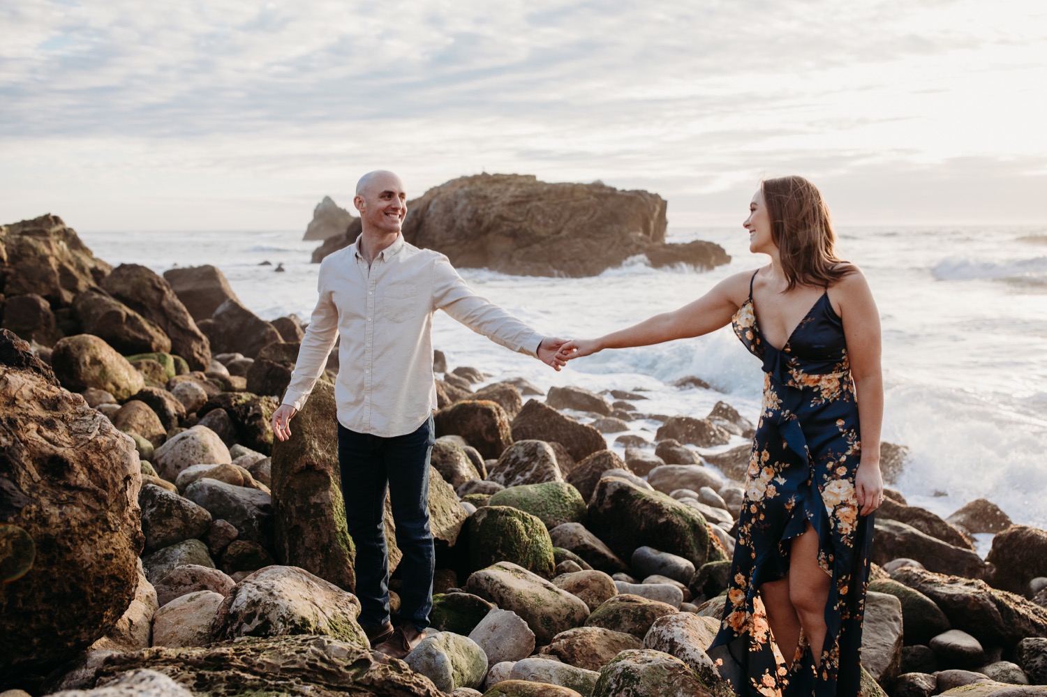 Couple reaches for each others hands while standing on a rocky beach at the Pacific Ocean during their San Francisco engagement photoshoot.
