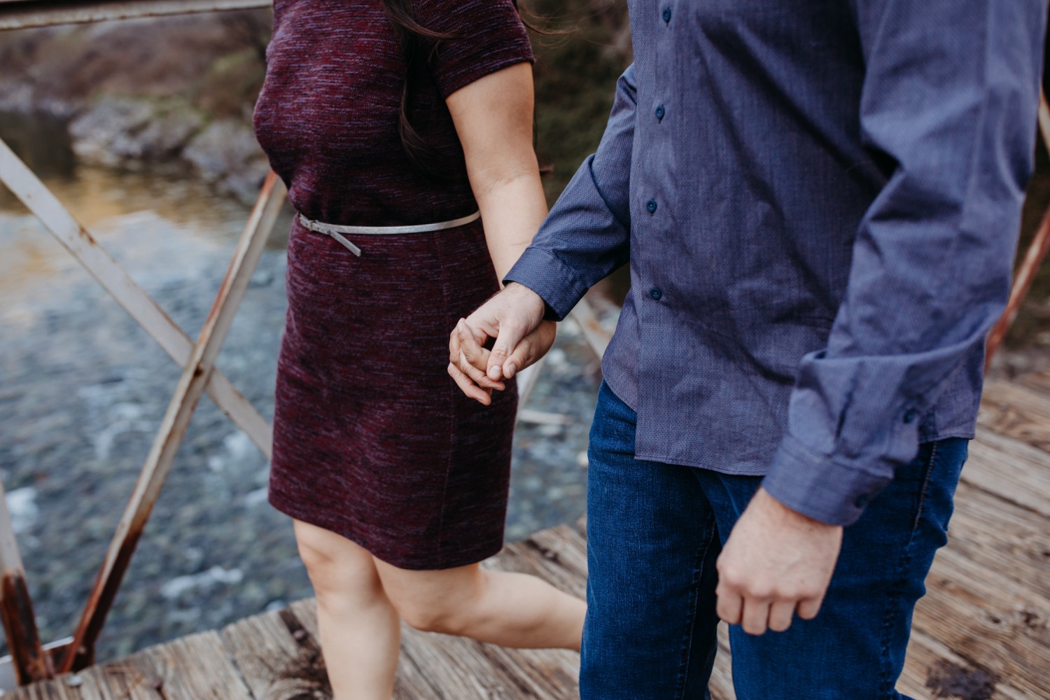 Couple holding hands as they cross a suspension bridge over a river on their engagement photoshoot.