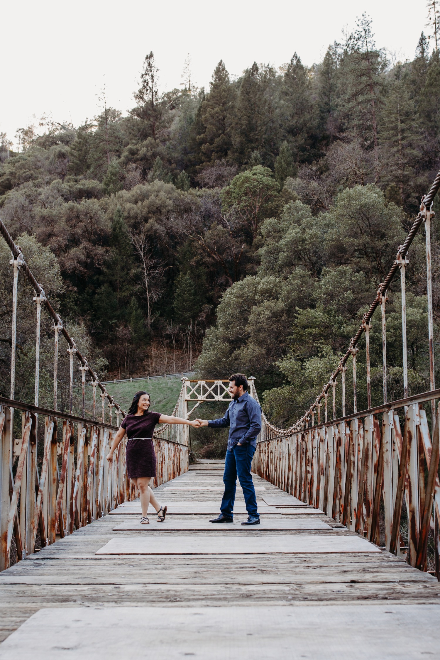 Couple dances on a suspension bridge with green forest in the distance during their American River engagement photoshoot.
