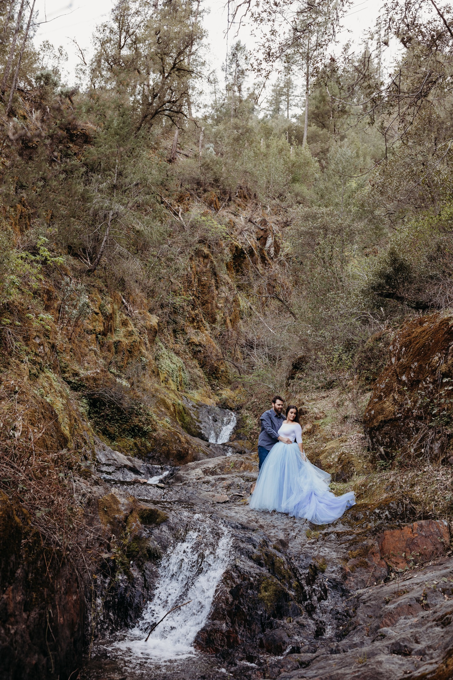 Couple stands in an embrace above a waterfall with the woman in a large light blue tulle skirt during their American River engagement photoshoot.