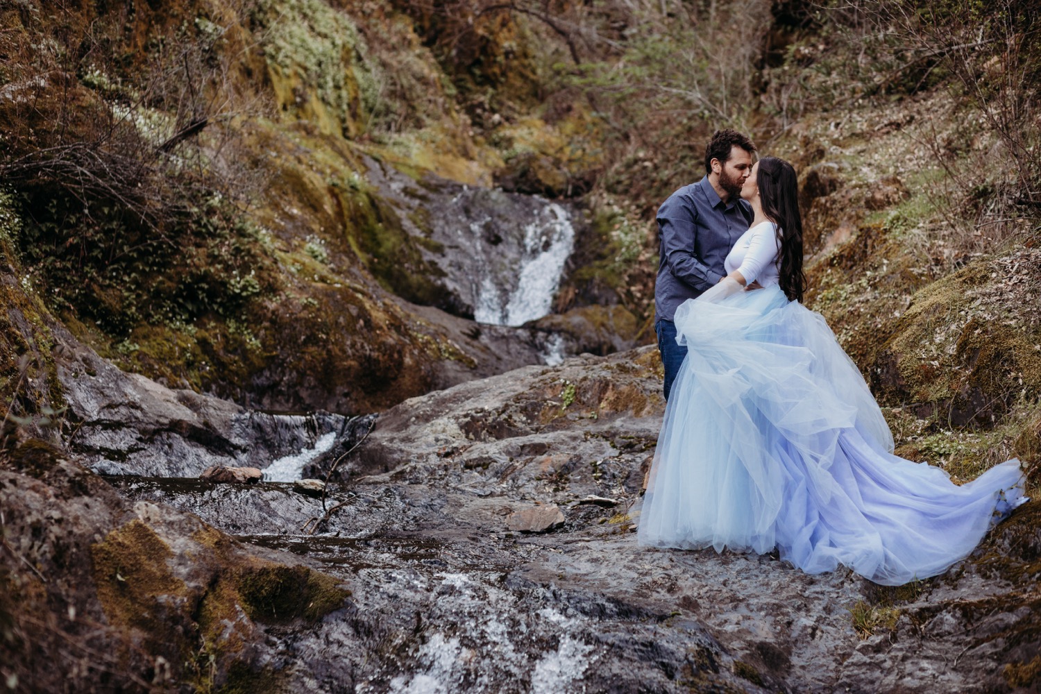 Couple kisses alongside a waterfall as the woman twirls her light blue tulle skirt.