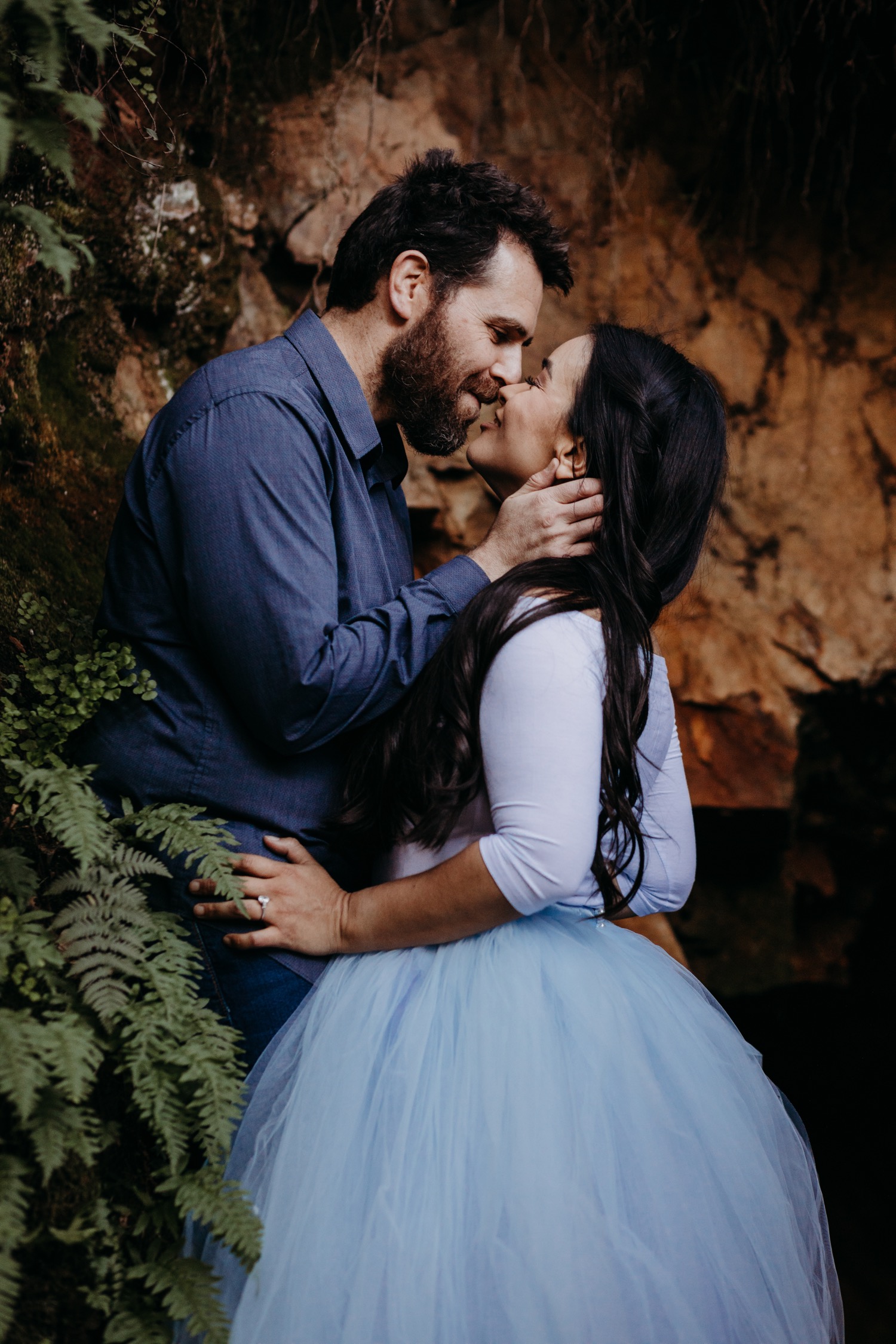 Man leads in to kiss his fiance amidst the greenery of the American River on their engagement photoshoot.