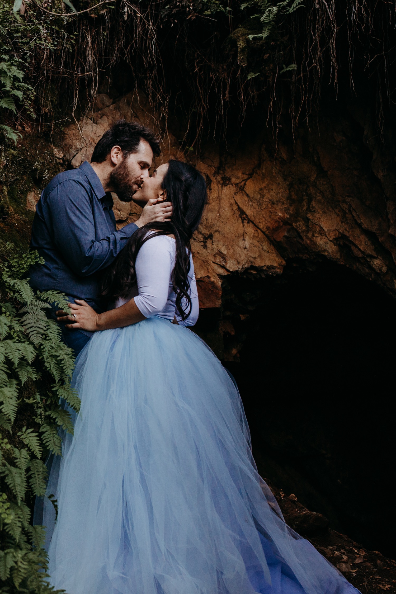 Couple kisses against rock wall with woman standing in light blue tulle skirt during their American River engagement photoshoot.