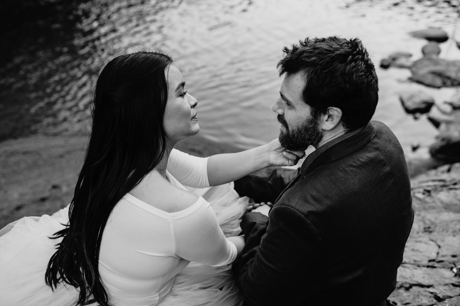 Woman holds her fiance's chin as they sit along side the American River on their engagement photoshoot.