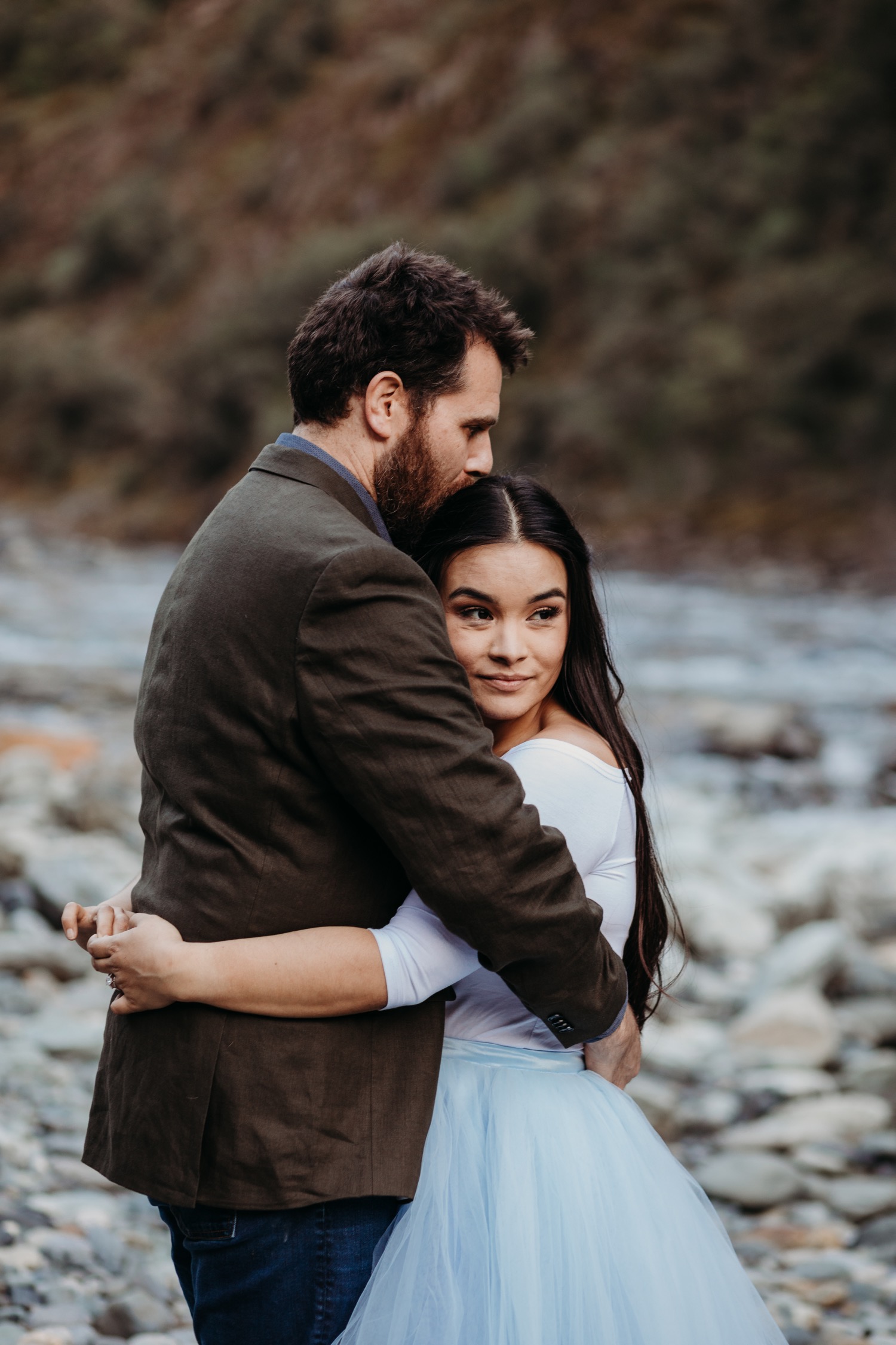 Couple embraces as the man kisses his fiance's head alongside the American River for their engagement photos.