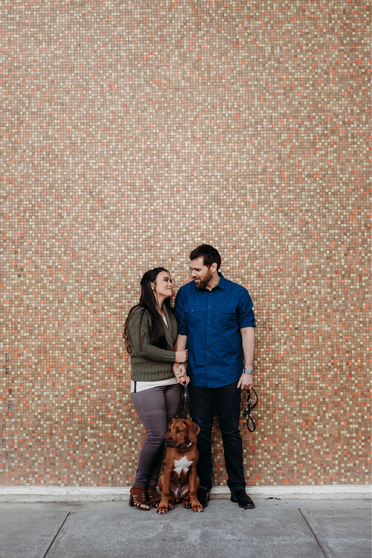 Couple stands gazing at each other with their dog in front of a pink tile wall during their Davis engagement photoshoot.