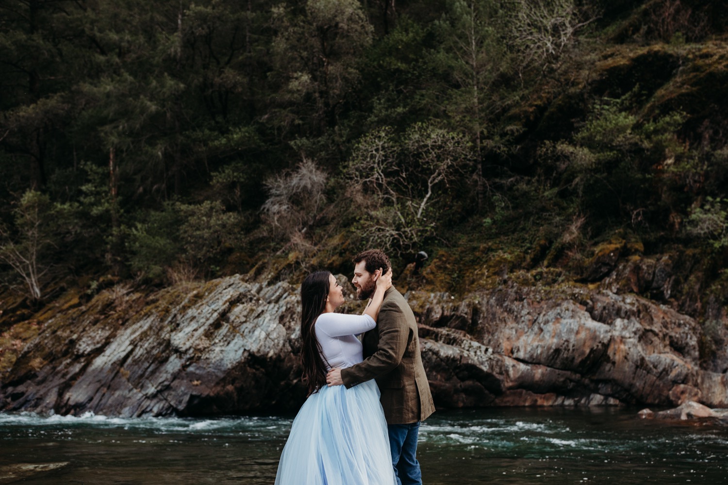 Couple stands in an embrace with the rushing American River in the background during their engagement photoshoot.