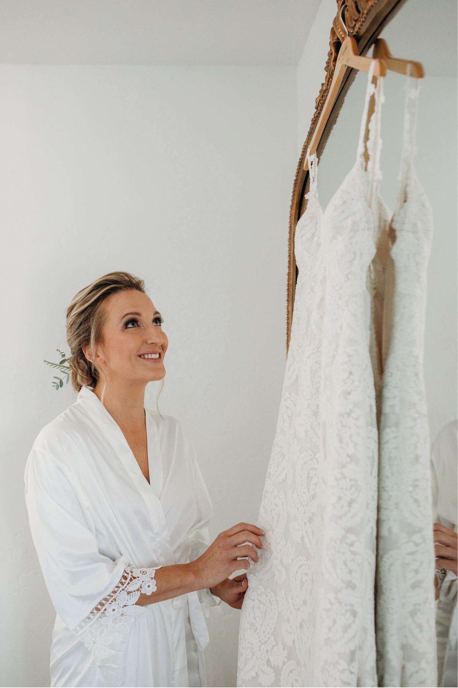Bride smiles while looking at her white lace wedding dress that's hanging on a wood hanger in front of a mirror. Liz Koston Photography.