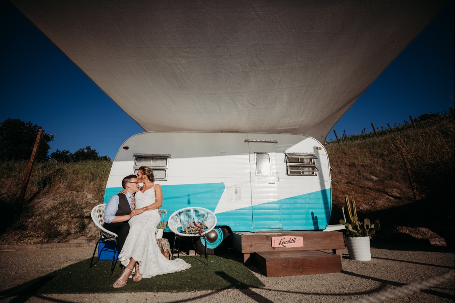 Brides sit on a chair in front of a small blue and white trailer before their Paso Robles wedding reception. Liz Koston Photography.