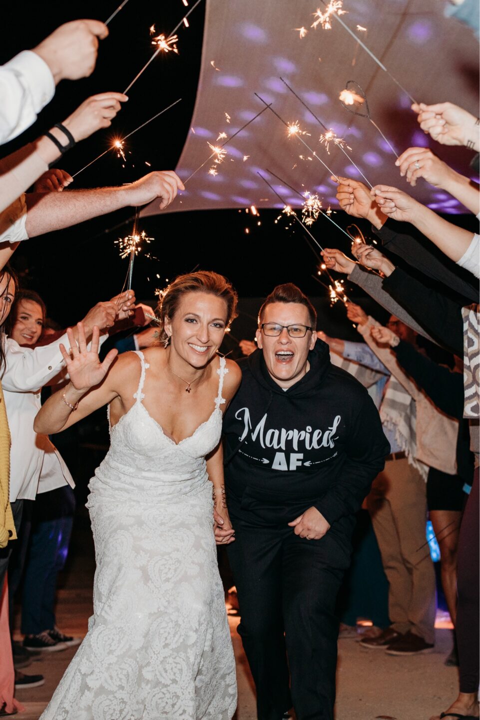 Brides walk out of their wedding reception under sparklers, one in a white lace wedding dress and the other in a sweatshirt that says "Married AF". Liz Koston Photography. 