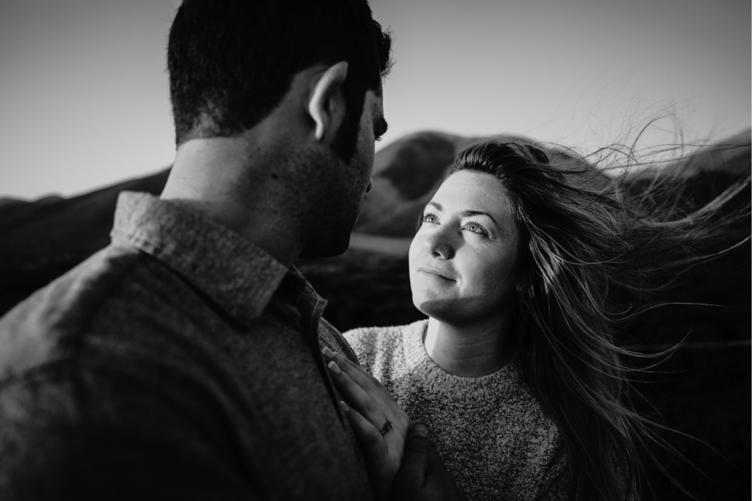 Black and white image of couple gazing at each other lovingly as the wind whips the woman's hair during their Big Sur Engagement photo session.