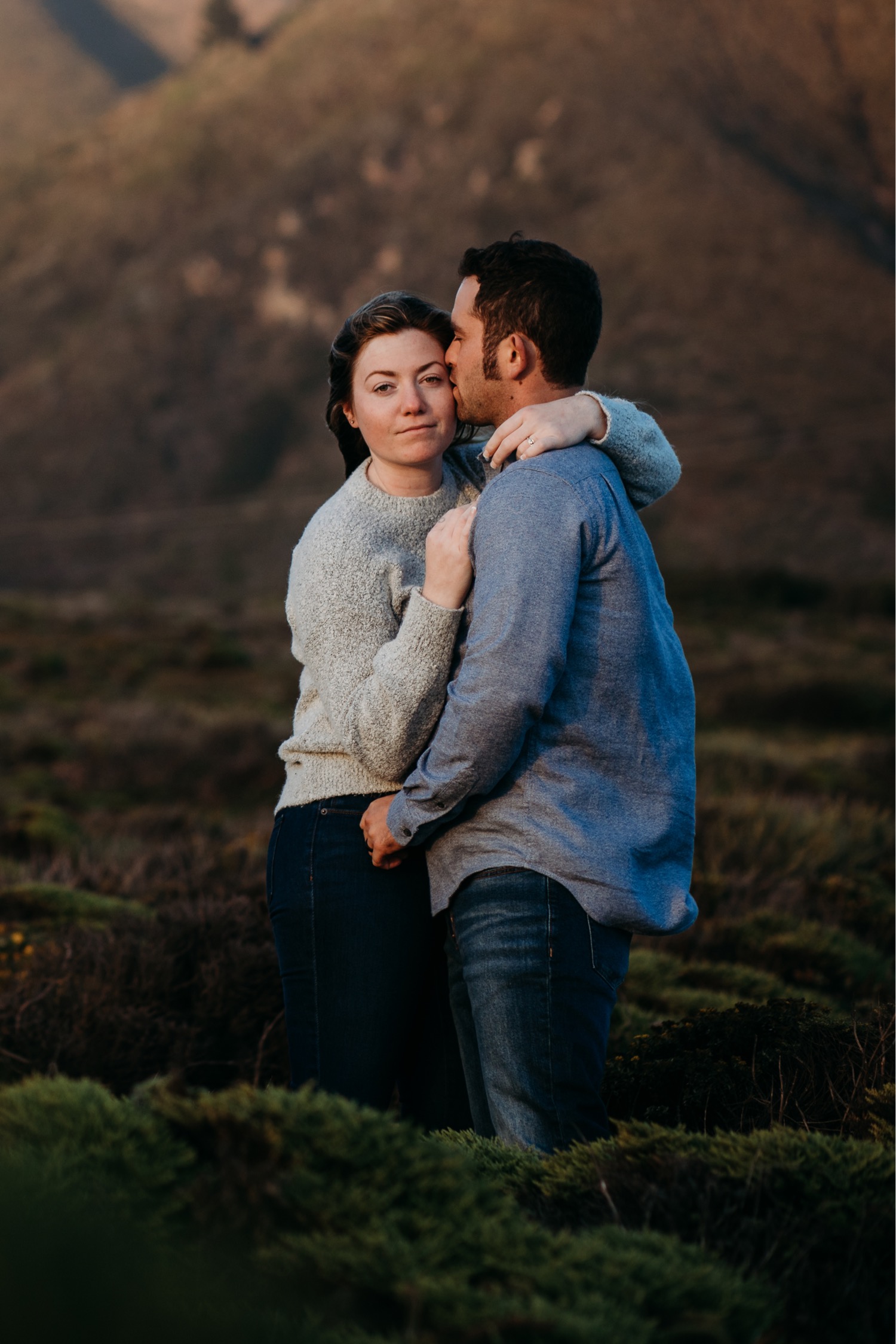 Couple embraces during their engagement photoshoot in Big Sur, California. The woman gazes at the camera as the man kisses her forehead.
