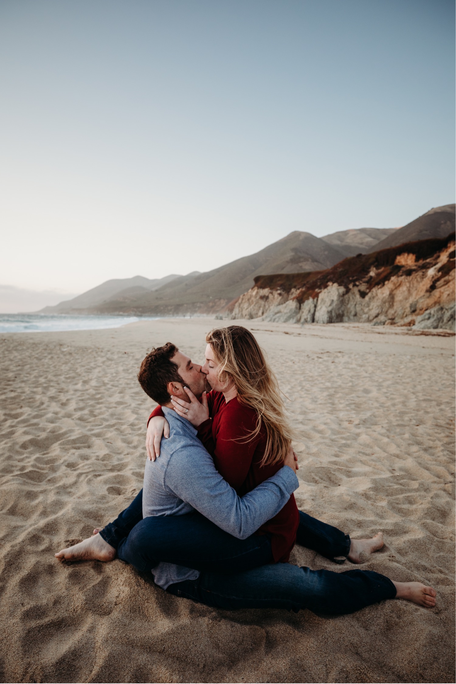 Couple sits in sand on the beach in Big Sur kissing each other passionately on their Big Sur, California engagement photoshoot.