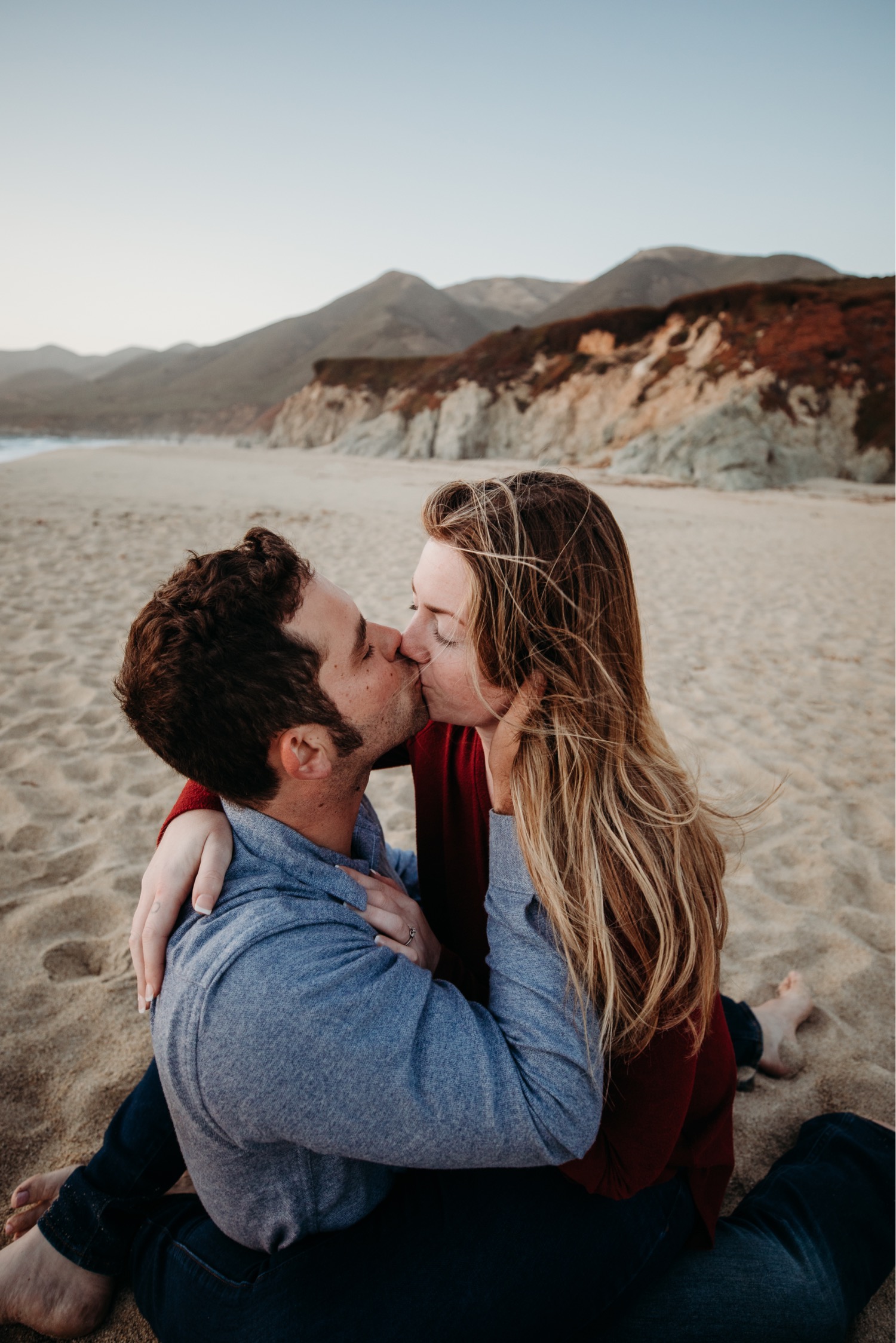 Couple sits in sand on the beach in Big Sur kissing each other passionately.