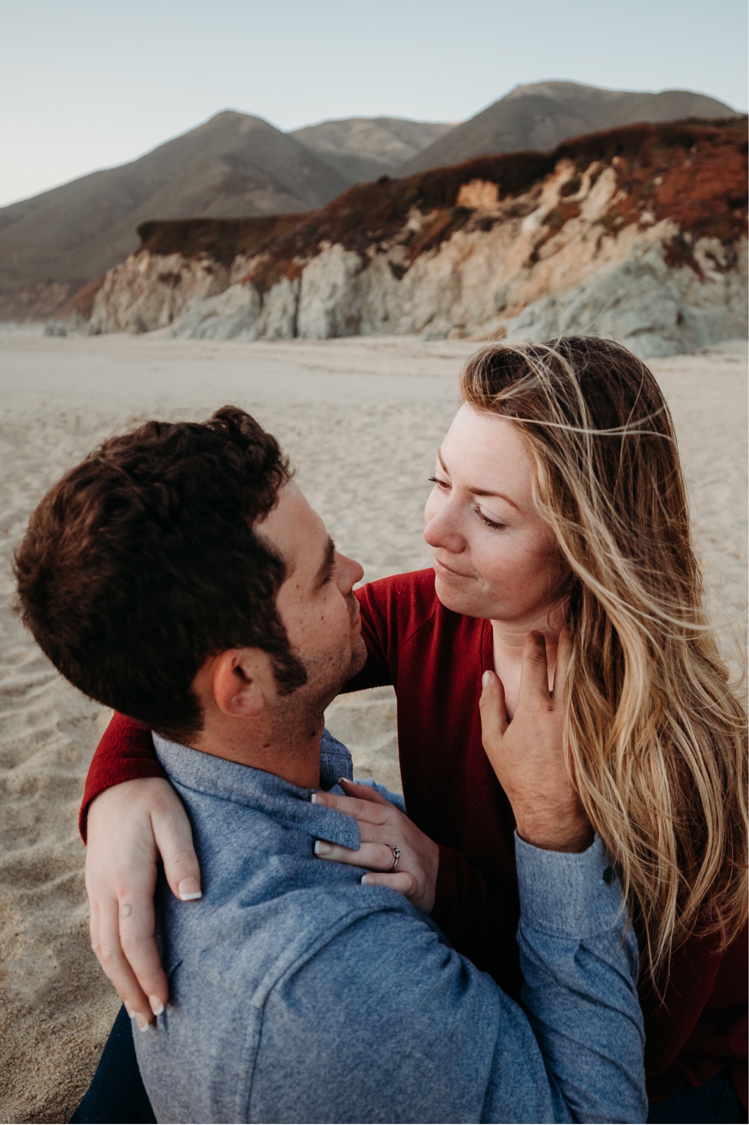 Couple sits in the sand on the beach in Big Sur gazing at each other lovingly as the man brushes the woman's hair from her face. 