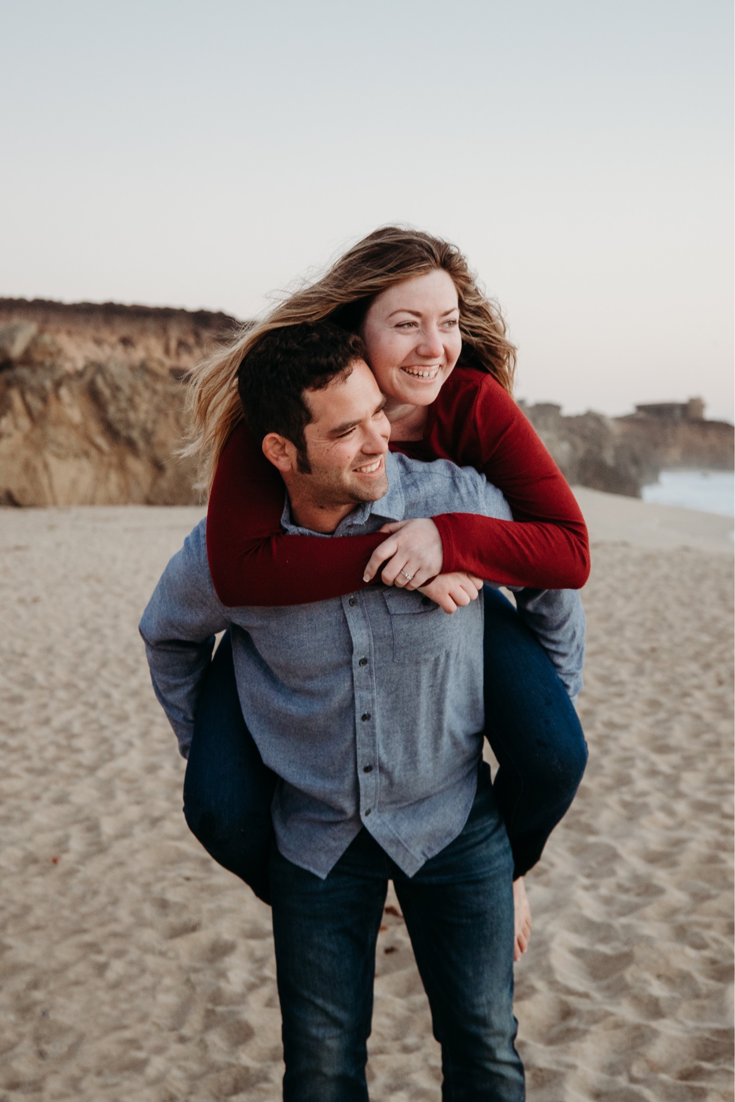 Man gives woman a piggy back ride on the beach in Big Sur as they both look towards the ocean.