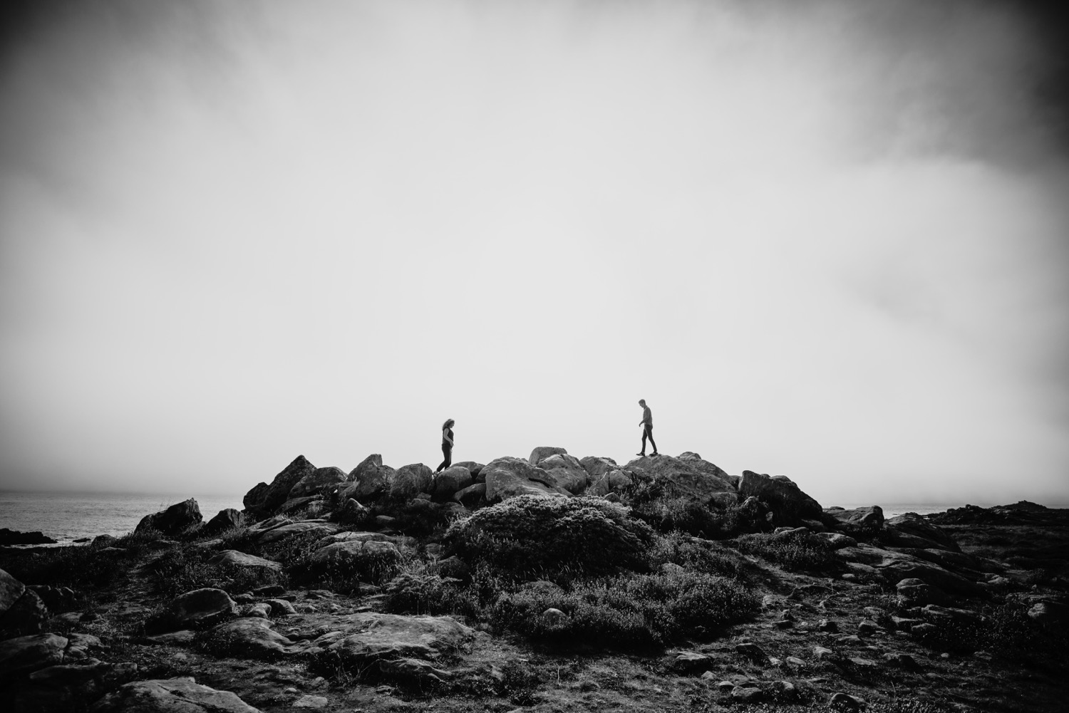 Black and white image of a couple walking towards each other over large boulders in Salt Point Park, CA