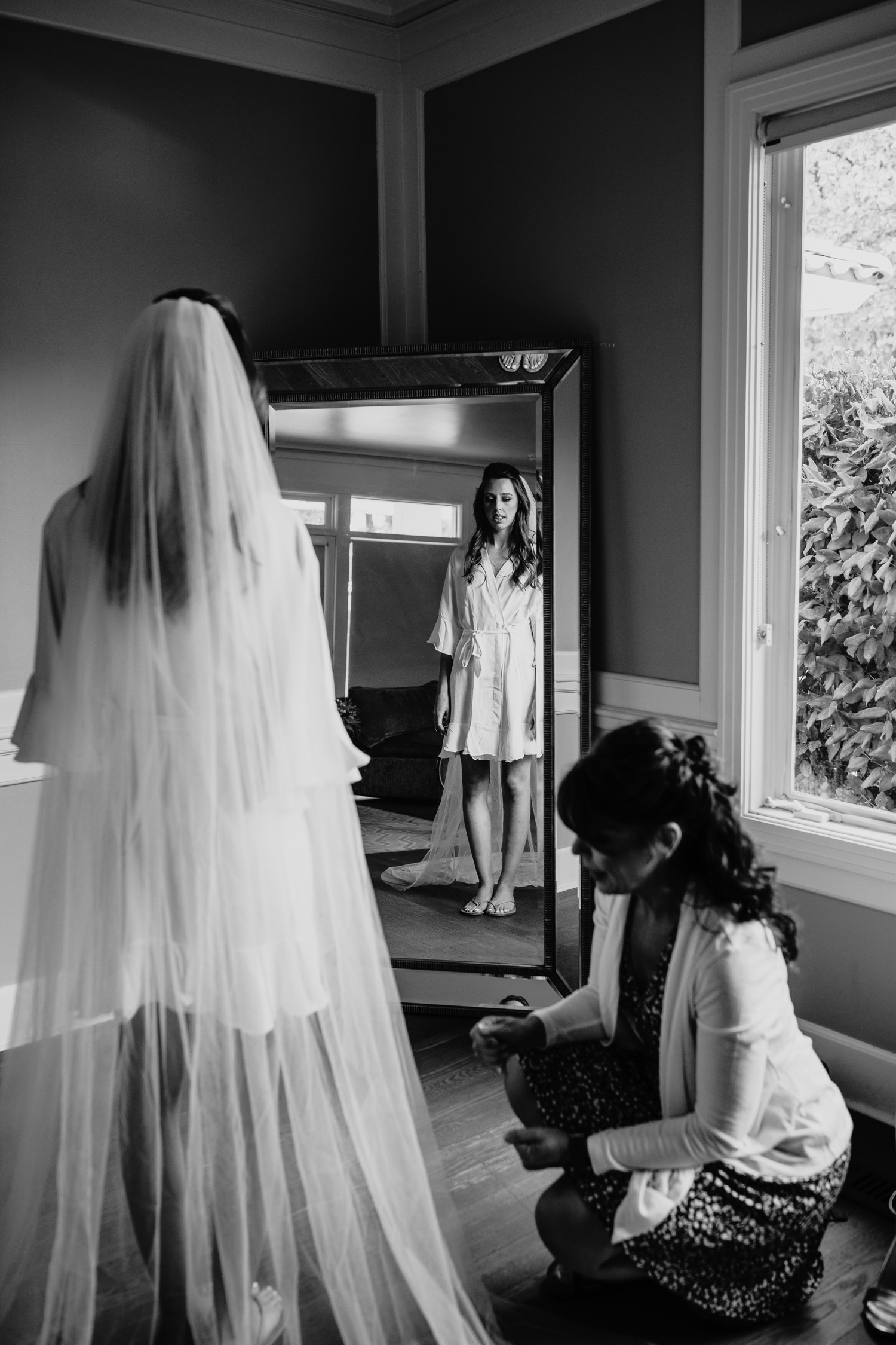 Bride standing in her veil and robe looking in the mirror as she gets ready for her wedding at the Maples in Woodland, CA.